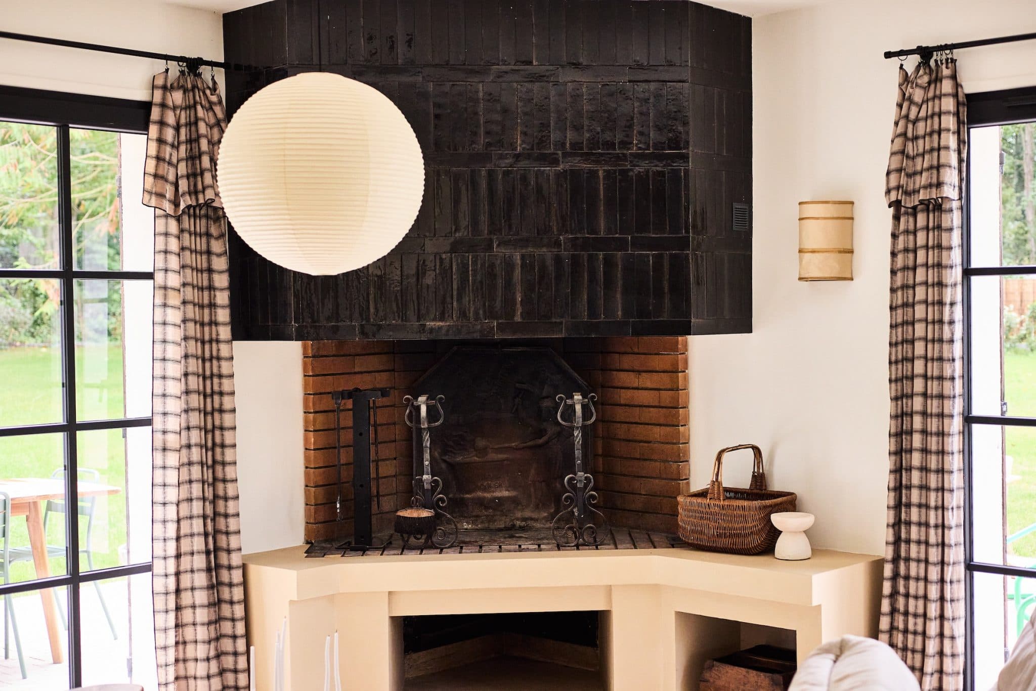 A cozy living room features a unique corner fireplace with black tiles, surrounded by neutral walls. There's a woven basket over the fireplace and minimalist decor. Two large windows with checkered curtains offer natural light and views of the greenery outside.