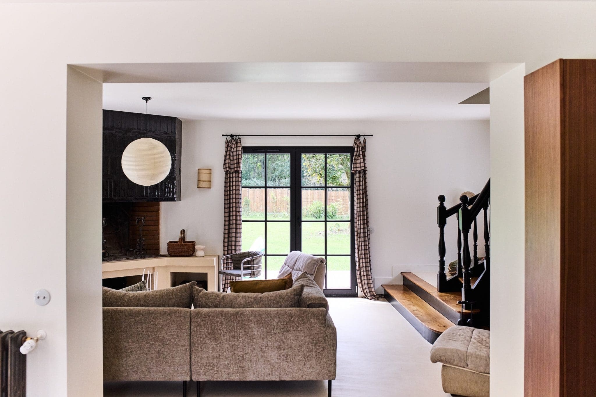 A comfortable living room with a gray corner sofa facing a fireplace. The room features large windows with patterned curtains, a spherical hanging lamp and a garden view. Wooden stairs with a black banister lead to another space.