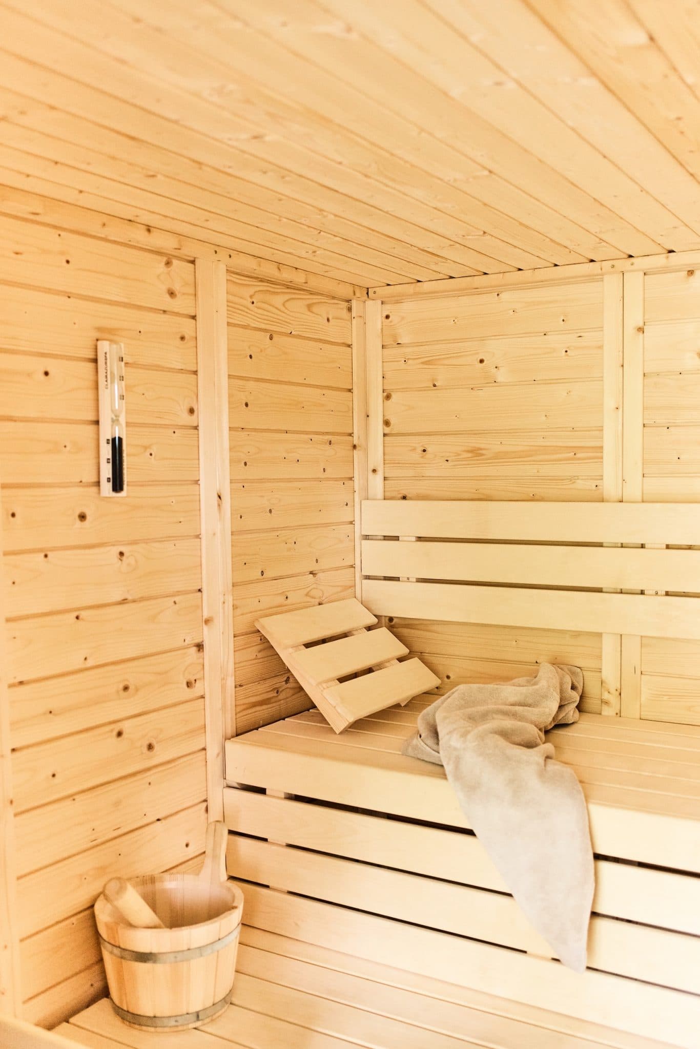 Wooden sauna interior with light-colored walls and ceiling. A wooden bench is covered with a soft towel. A small wooden bucket sits on the floor and a thermometer is fixed to the wall, underlining a warm, welcoming atmosphere.