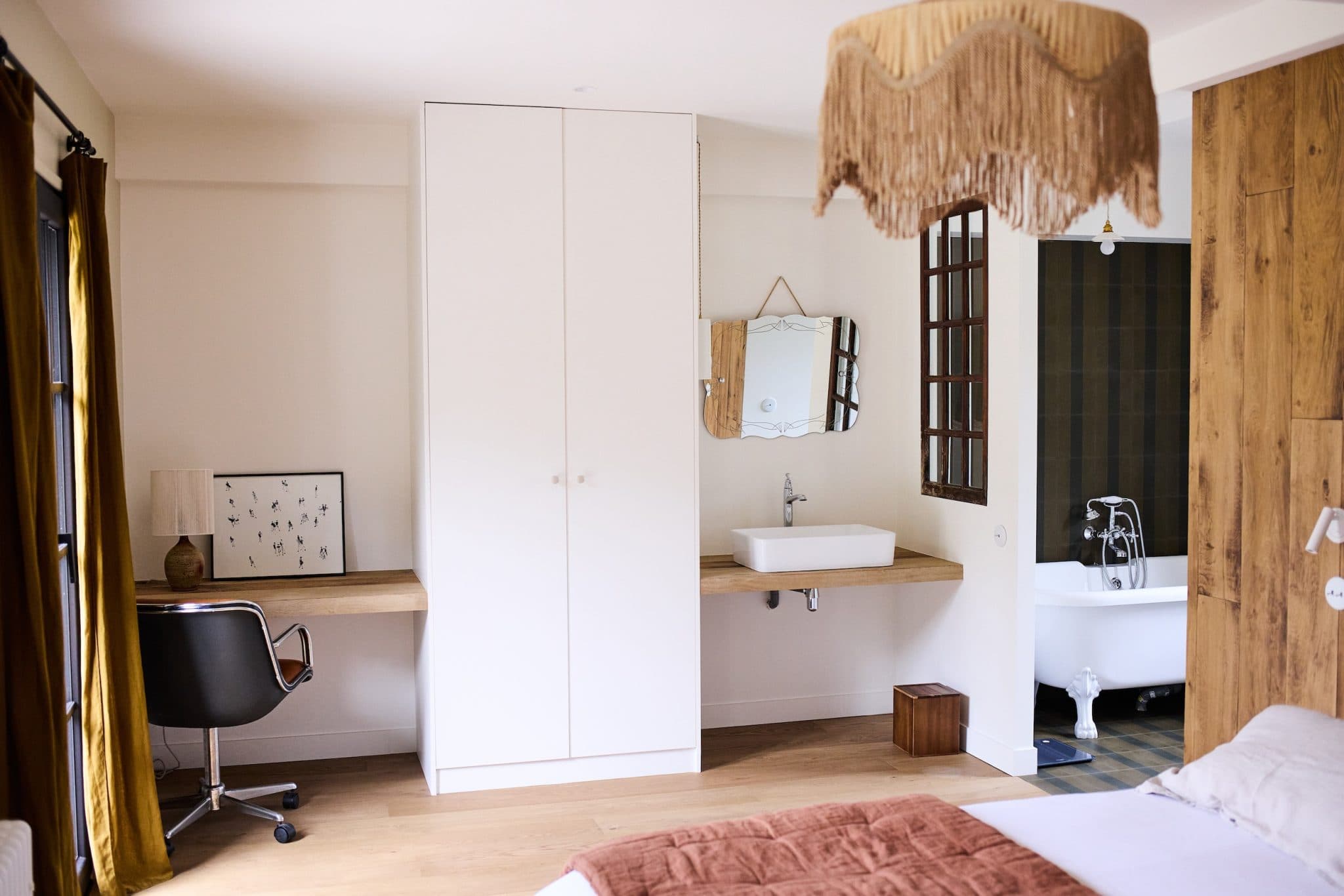 A comfortable bedroom with a white wardrobe, a wooden desk with chair and a wall-mounted washbasin with mirror. A freestanding bathtub is visible through an open door. The room features warm tones and a fringed ceiling light.