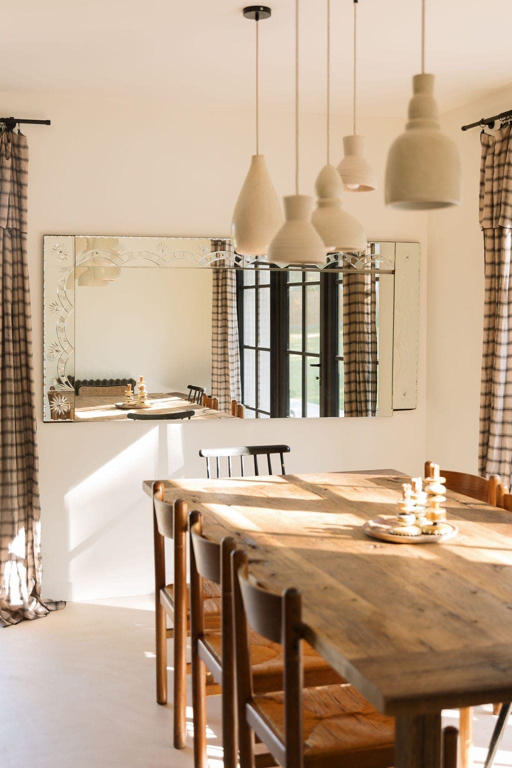 A dining room with a wooden table and chairs, decorated with a checkered table runner, lit by hanging lights. A large, ornate mirror reflects natural light from the windows, and checkered curtains frame the window.