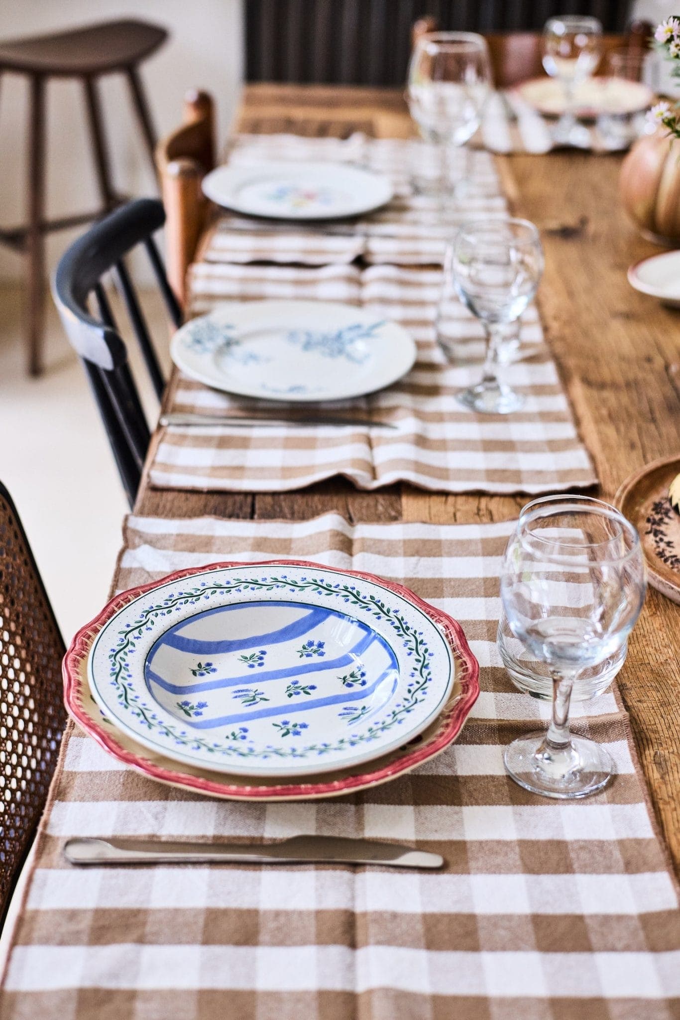 A dining table is set with checkered tablecloths, white and blue floral plates, clear wine glasses and cutlery. The table extends diagonally across the picture, creating a warm, welcoming atmosphere.