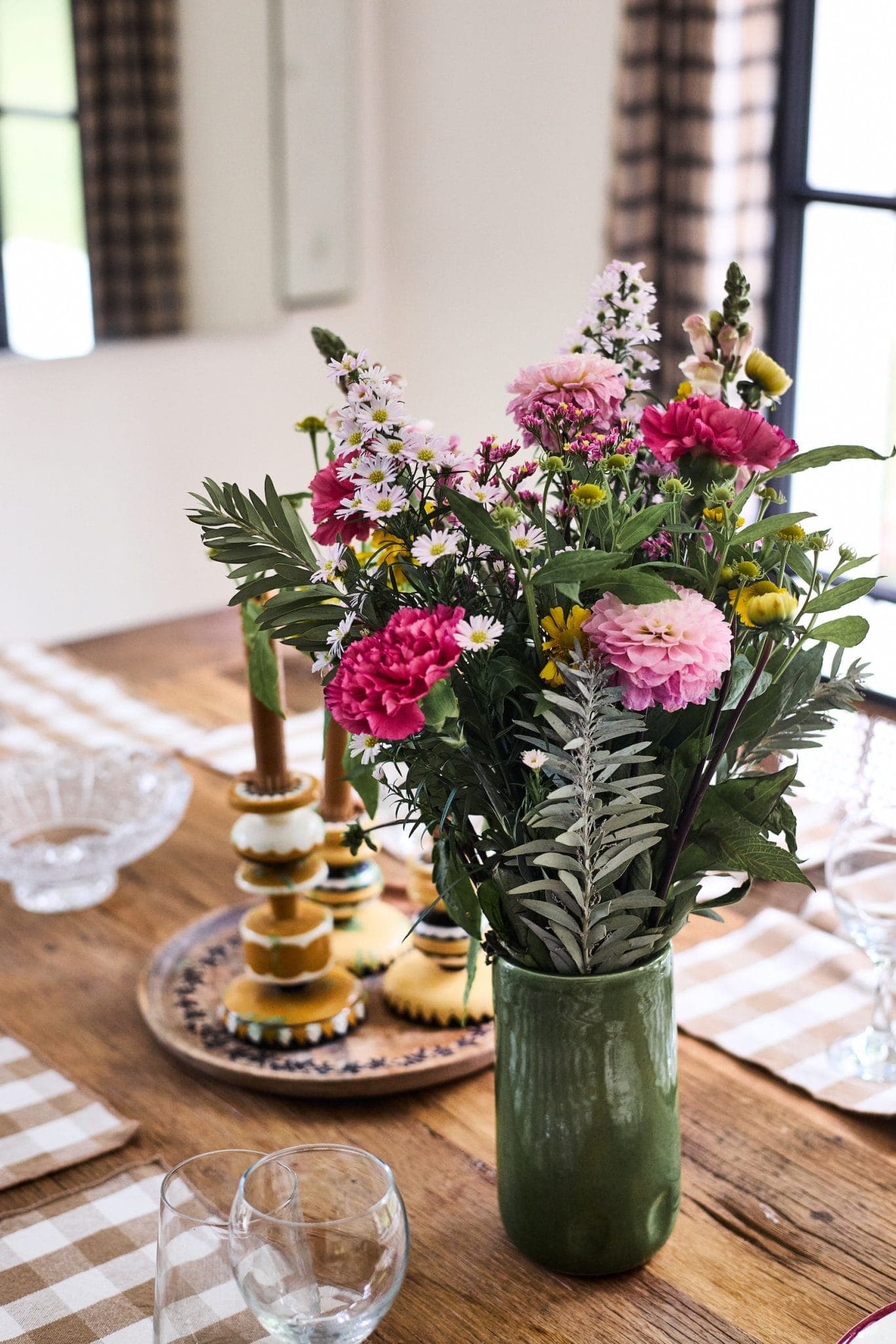 A green vase with a bouquet of pink and purple flowers sits on a wooden dining table. There are checkered placemats, clear glassware and a wooden tray with decorative objects in the background.
