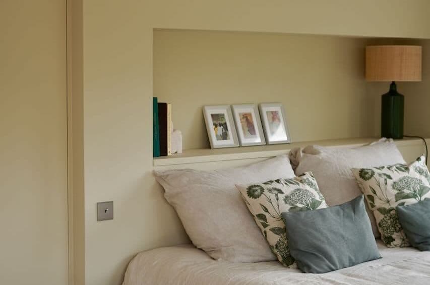 Une chambre confortable dispose d'un lit soigneusement fait avec des oreillers blancs et fleuris. Les accents en bois comprennent une petite table d'appoint bleue et une lampe. Trois photos encadrées sont exposées sur une étagère derrière le lit, avec une poutre en bois peinte au-dessus.