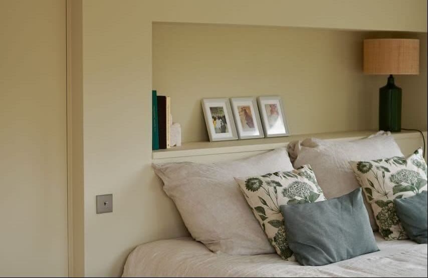 A comfortable bedroom features a carefully made bed with white and floral pillows. Wooden accents include a small blue side table and a lamp. Three framed photos are displayed on a shelf behind the bed, with a painted wooden beam above.