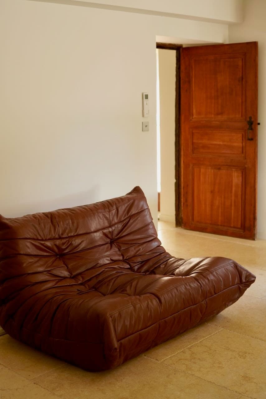A brown leather sofa rests on a light-colored floor in a minimalist room. The background is composed of a plain off-white wall and an open wooden door, creating a warm, welcoming atmosphere.