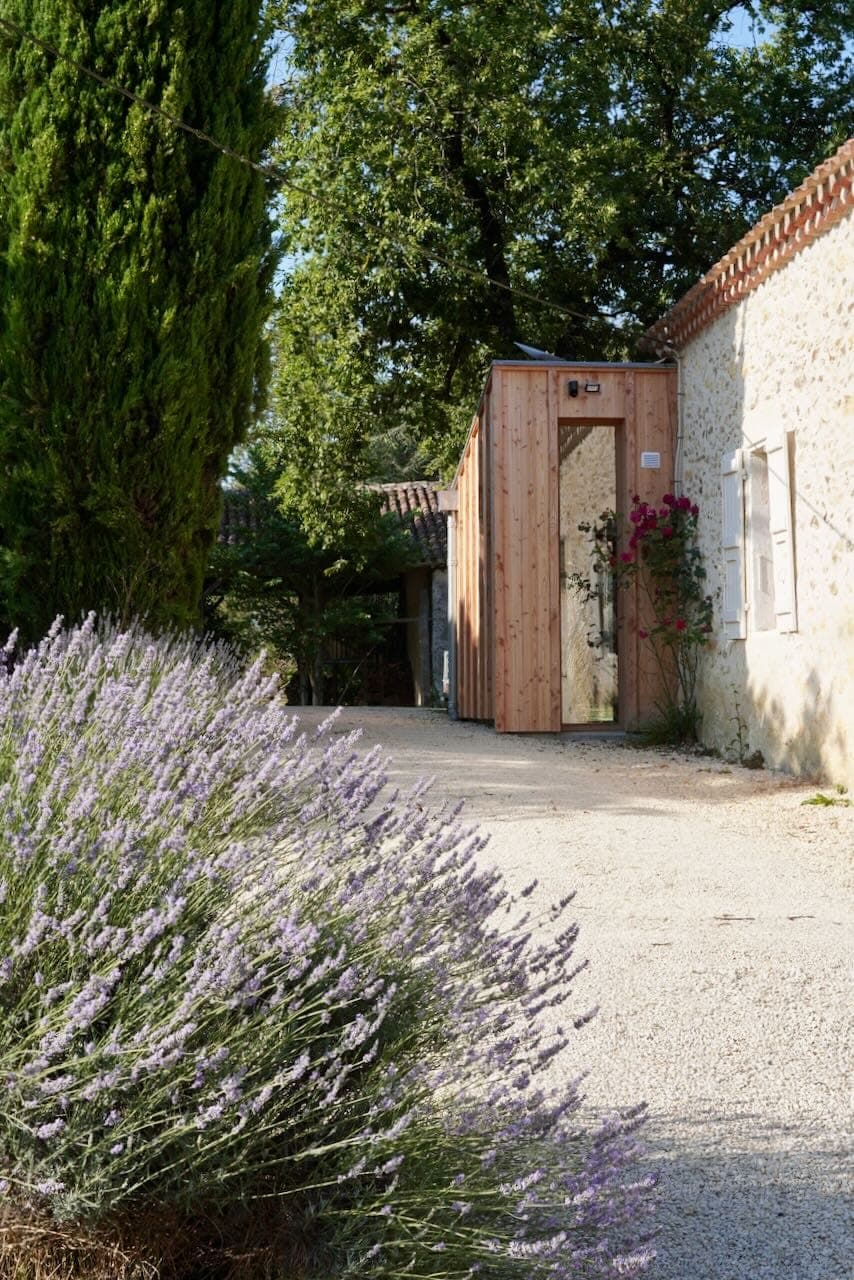 A picturesque rural scene with lavender in bloom in the foreground, a gravel path leading to a rustic stone building with a small wooden extension, surrounded by lush greenery and trees under a clear sky.