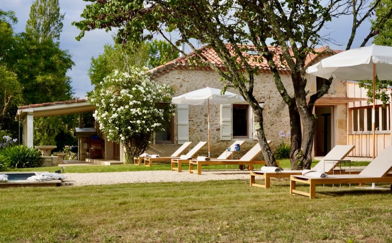 A rustic stone house with a red tile roof is surrounded by trees and green grass. White sun loungers and parasols are placed next to a small pool in the courtyard, creating a serene and welcoming outdoor setting.