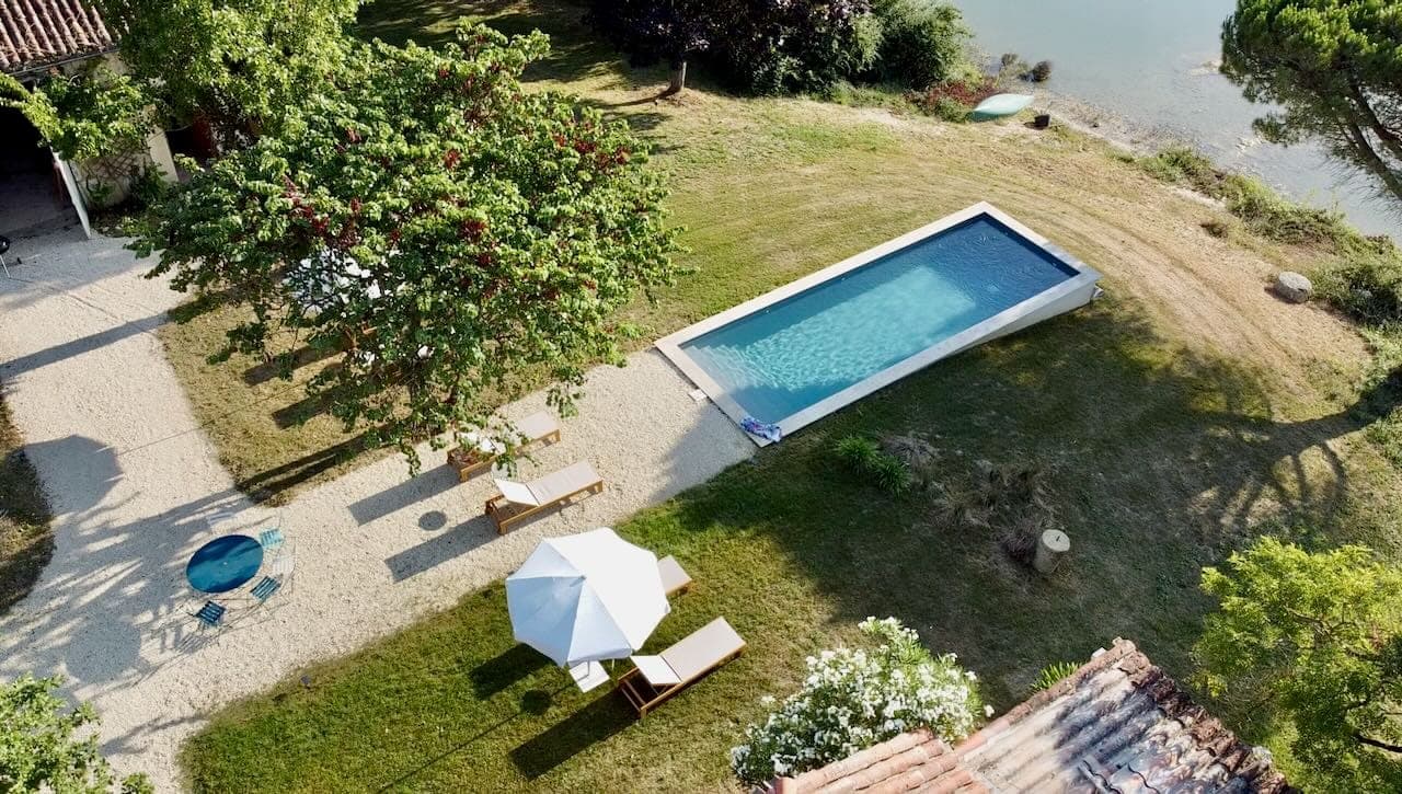 Aerial view of a quiet backyard with a rectangular swimming pool, surrounded by green grass and trees. Two sun loungers with a white parasol are on a nearby path, next to a small round table. A calm stretch of water is visible in the background.