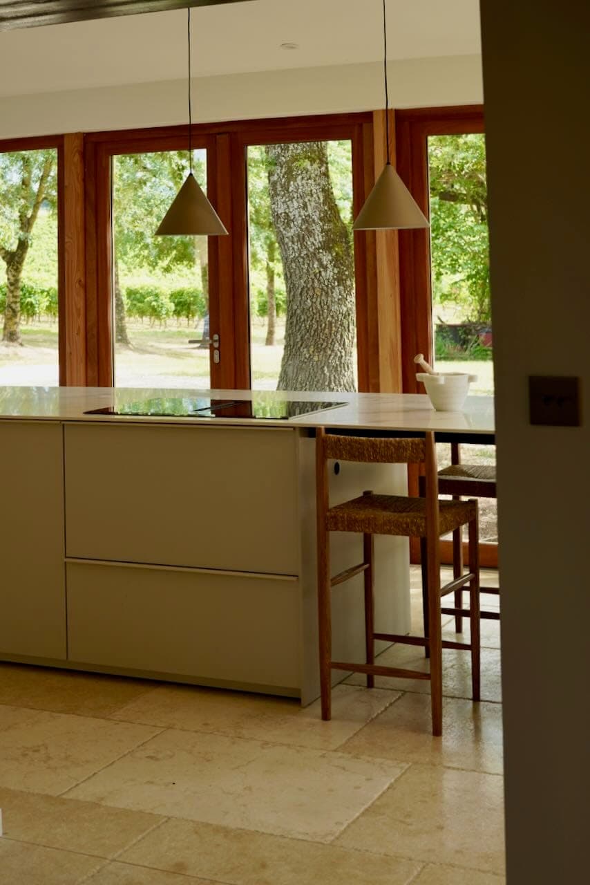 Modern kitchen with minimalist design, central island, two wooden chairs, pendant lights and large windows overlooking the lush greenery outside. The room exudes a bright, airy atmosphere.