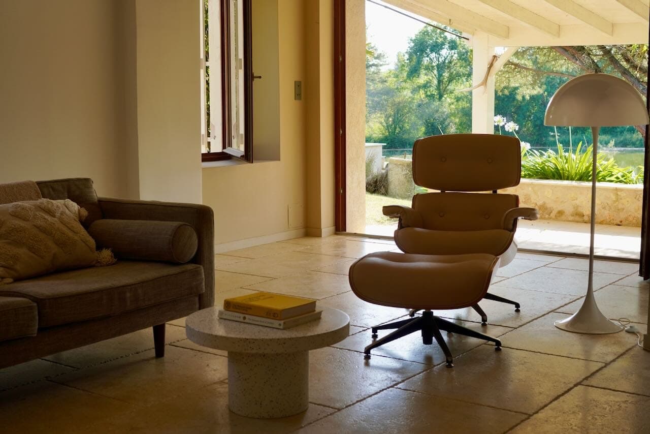 A comfortable living room with an open window letting in natural light. It includes a taupe sofa, an elegant chaise longue and footstool, a round coffee table with books and a large white floor lamp. A view of a lush garden can be admired from outside.
