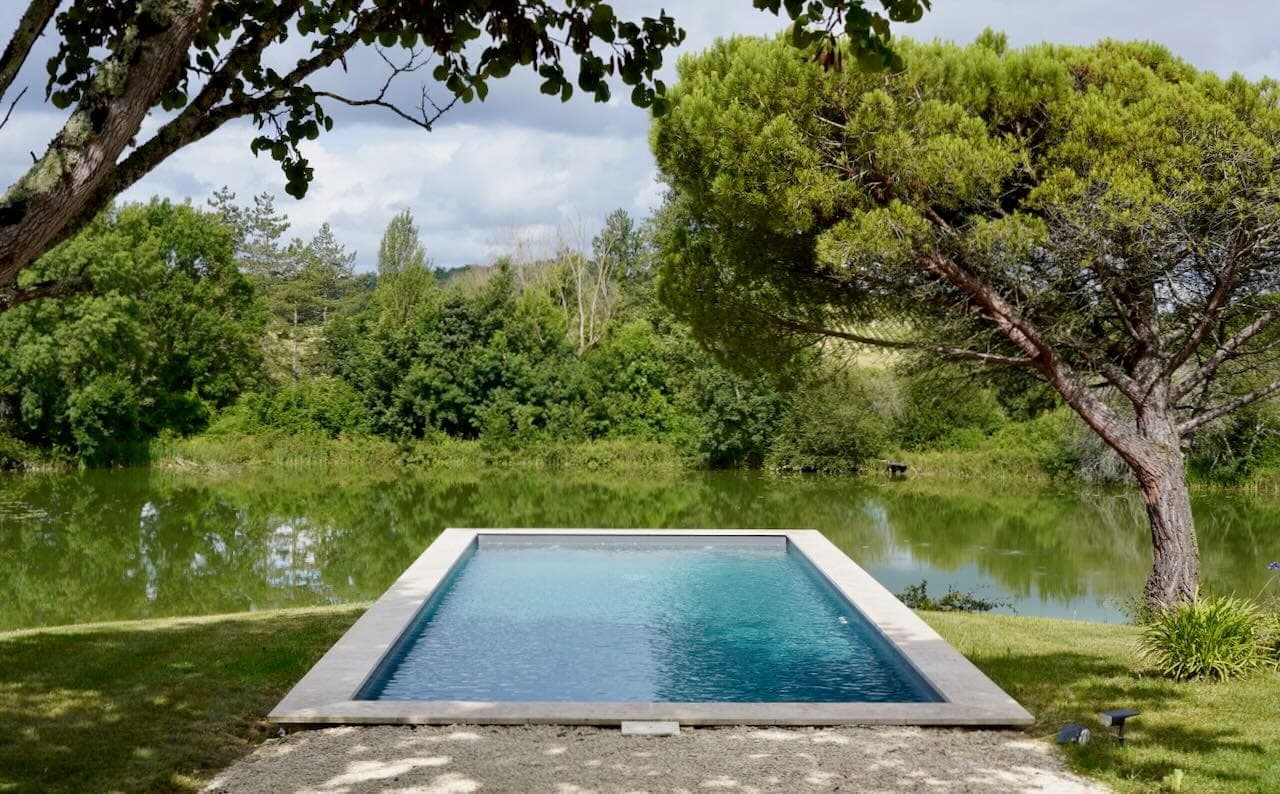 A rectangular infinity pool overlooks a serene body of water surrounded by lush green trees under cloudy skies. A large tree frames the right-hand side, creating a tranquil, welcoming outdoor setting.