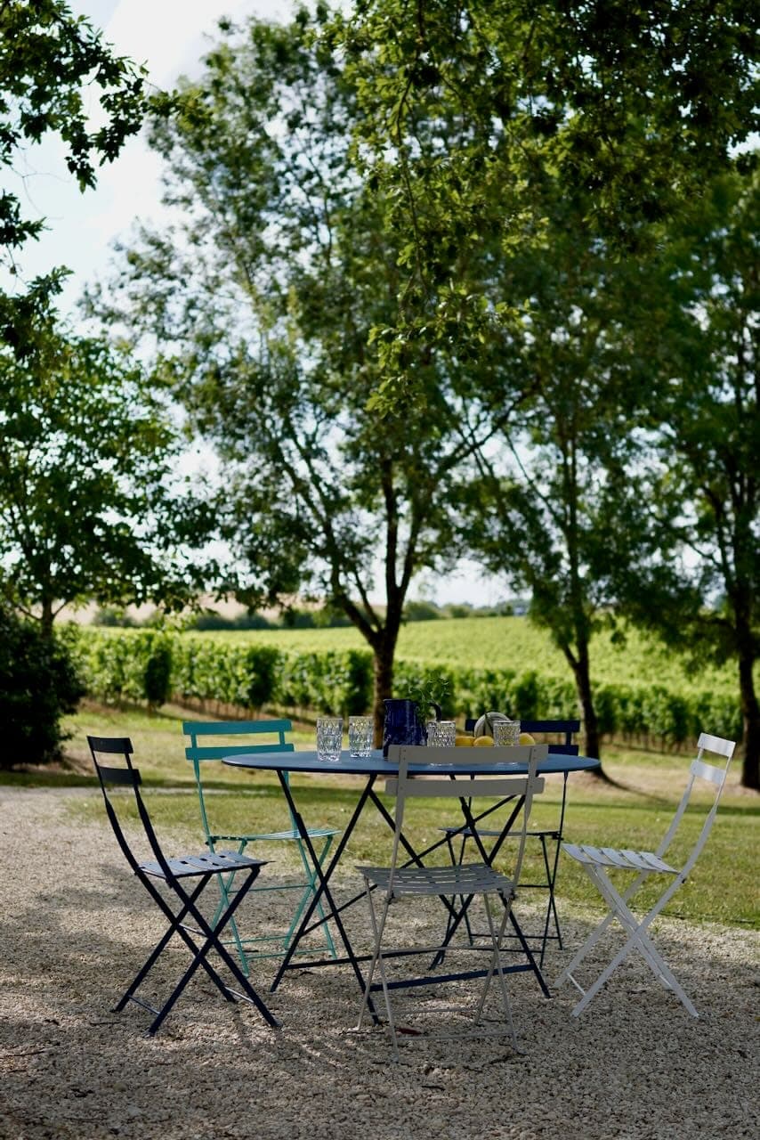 Folding chairs and a table with glasses are set in a gravelled area outside, surrounded by lush green trees and a vineyard in the background. The setting is peaceful and sunny.