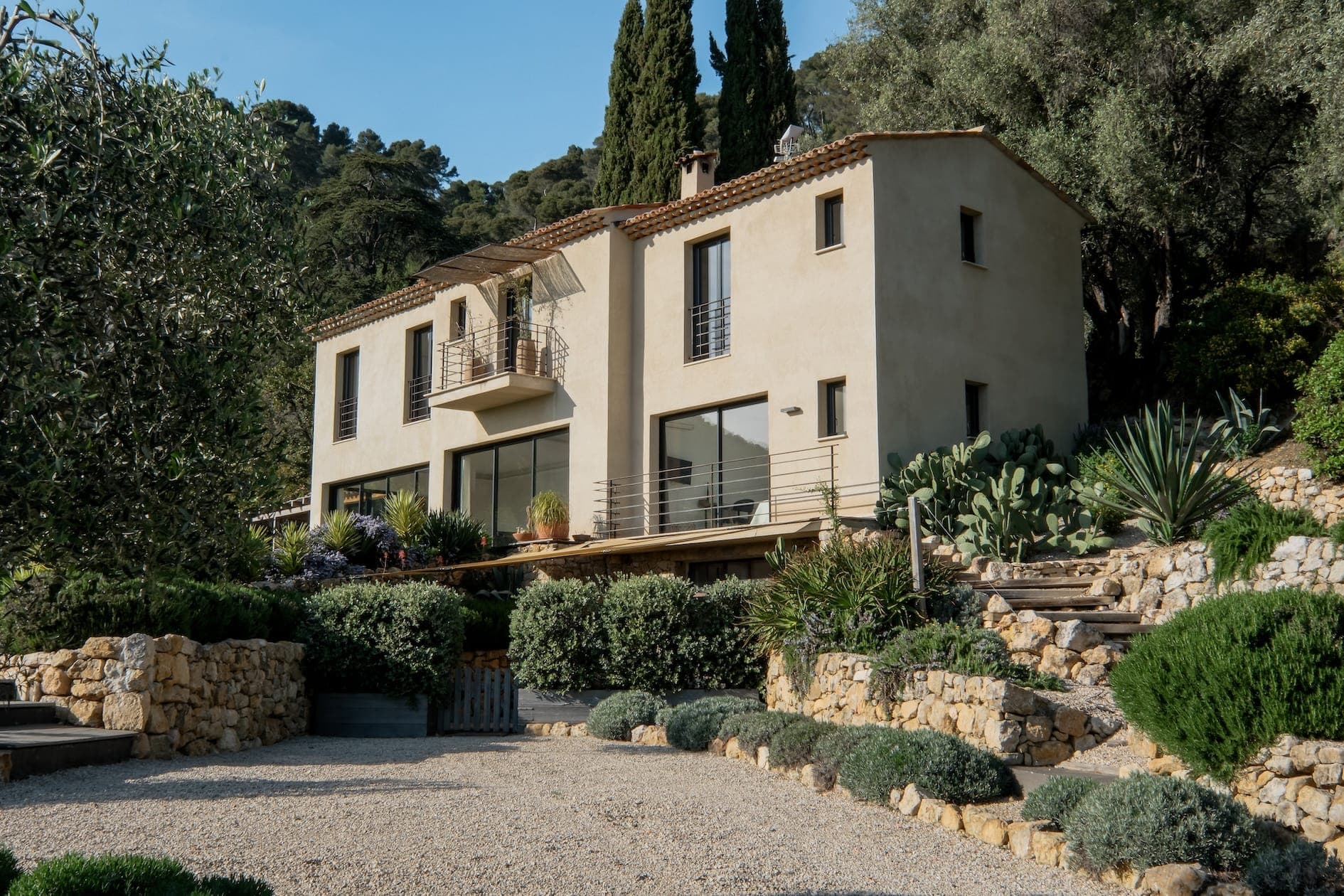 A Mediterranean-style house with light stucco walls and terracotta roof, surrounded by lush vegetation and stone landscaping. A gravel path leads to the entrance, and tall trees stand in the background under a clear sky.