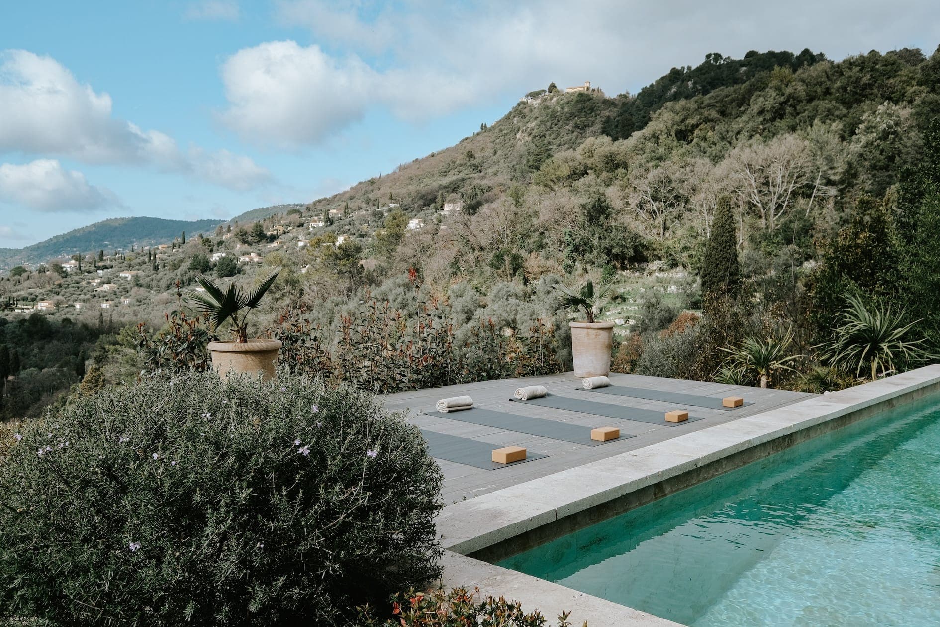 A serene outdoor yoga platform surrounded by lush greenery and mountains. Four yoga mats are neatly arranged with blocks and cushions. In the foreground, a turquoise pool sparkles and the sky above is partly cloudy.