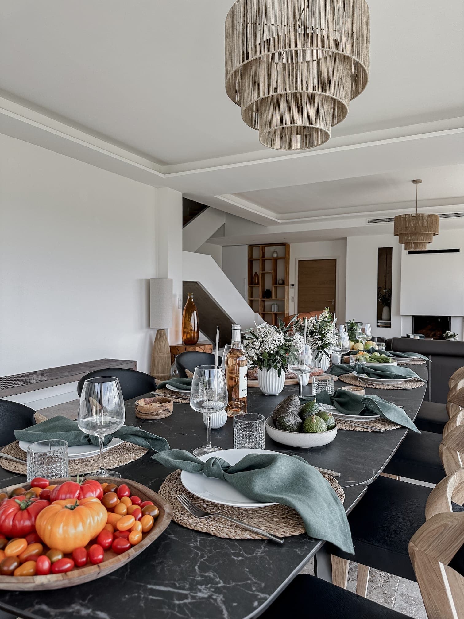 Elegant dining room with a long black table set for a meal. It includes wicker placemats, green napkins, wine glasses and matching dishes. Fresh produce and floral arrangements decorate the table. Modern decor in neutral tones and pendant lighting.