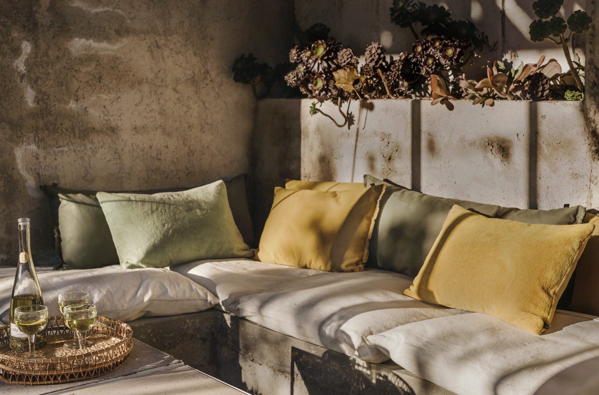 Cozy outdoor seating area with a corner sofa adorned with yellow and green cushions. Sunlight filters through, casting shadows on the beige wall and succulent plants above. A tray with wine glasses and a bottle sits on the table.