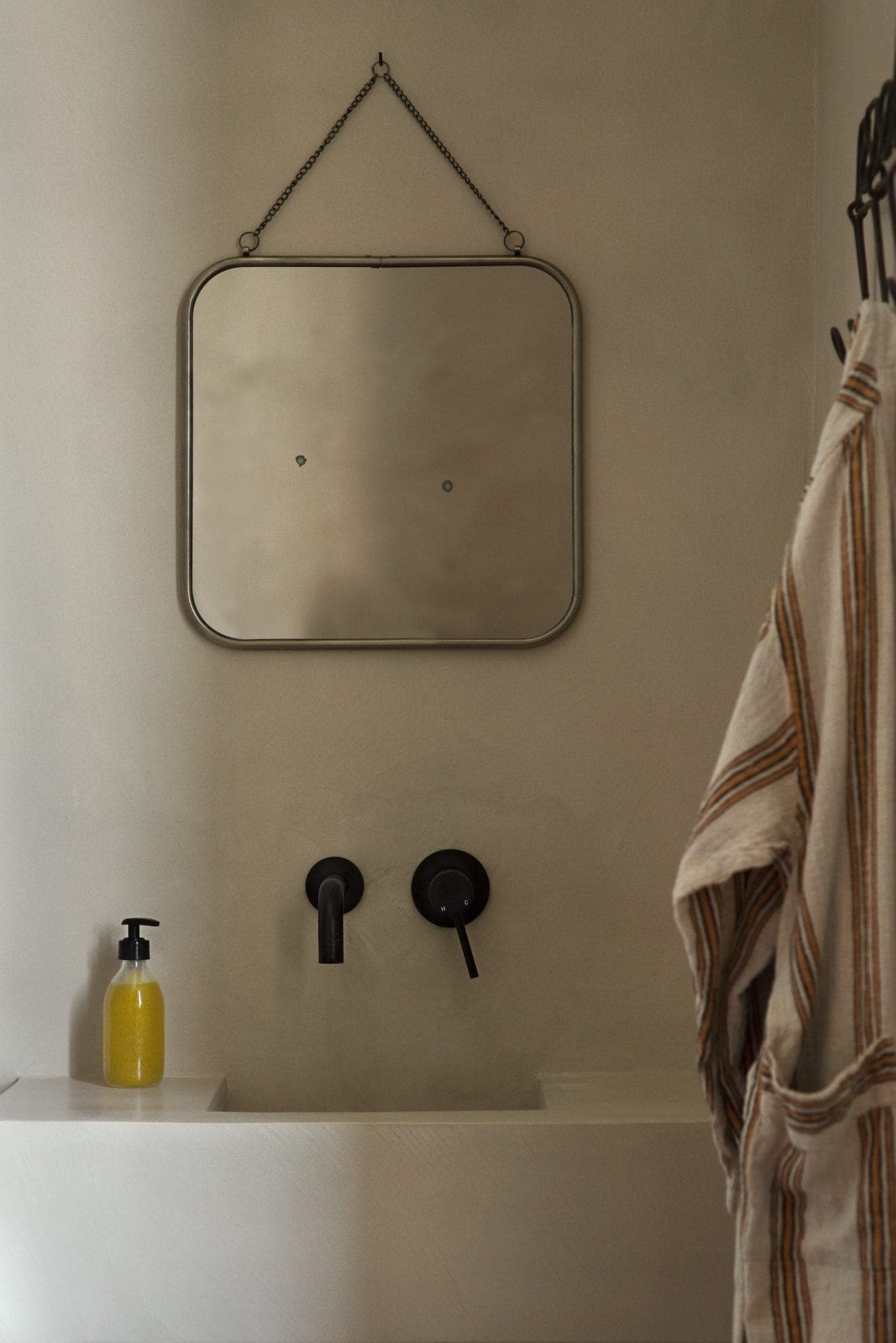 Minimalist bathroom interior with a square mirror hanging above a wall-mounted sink. A striped towel is visible on the right and a yellow soap dispenser is on the left corner of the sink. The overall color scheme is neutral and earthy.