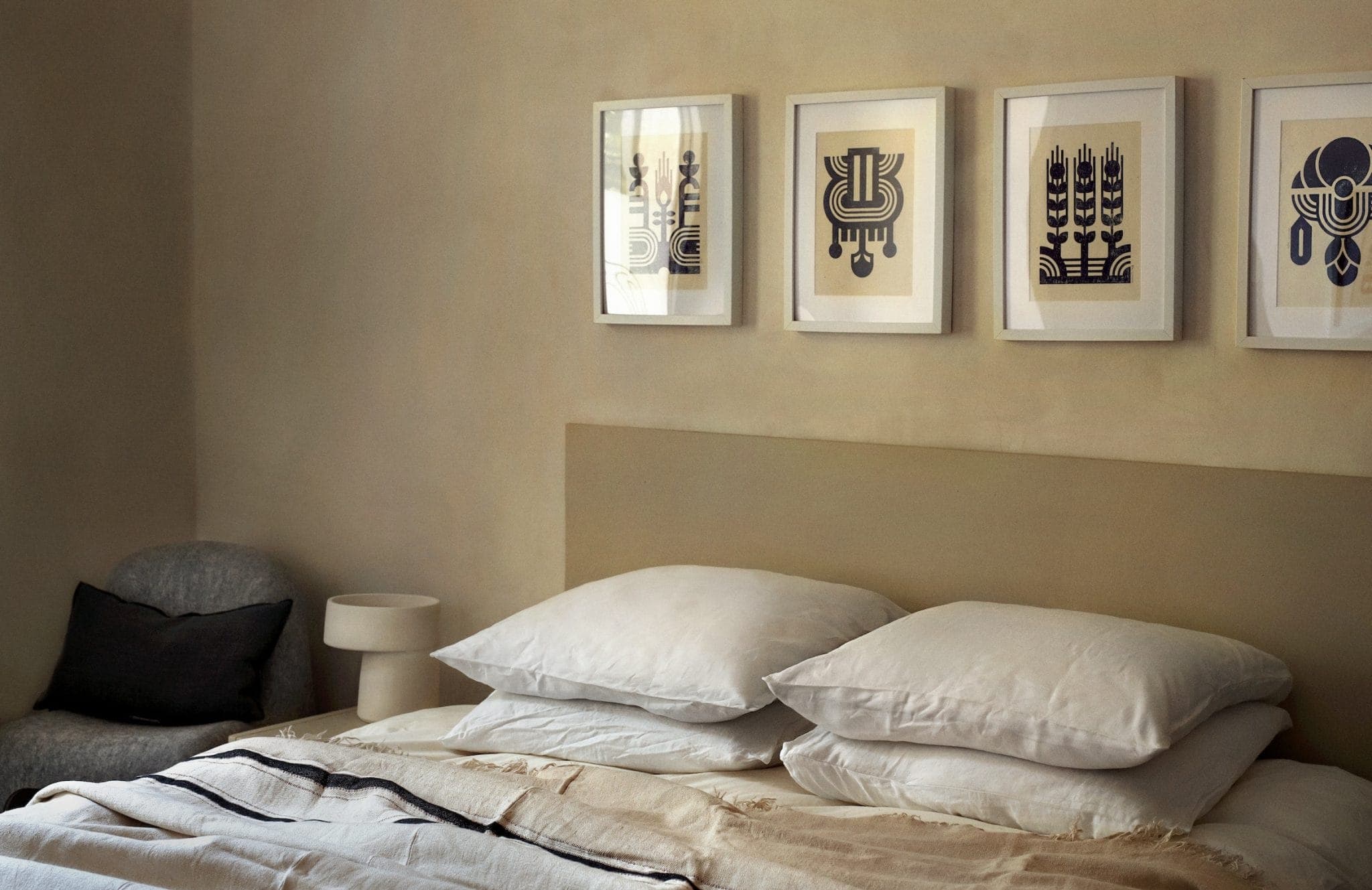 A cozy bedroom with a neatly made bed, featuring white pillows and a beige blanket. Above the bed, four abstract framed artworks in black and white hang on the wall. A small lamp and a chair are visible in the corner.