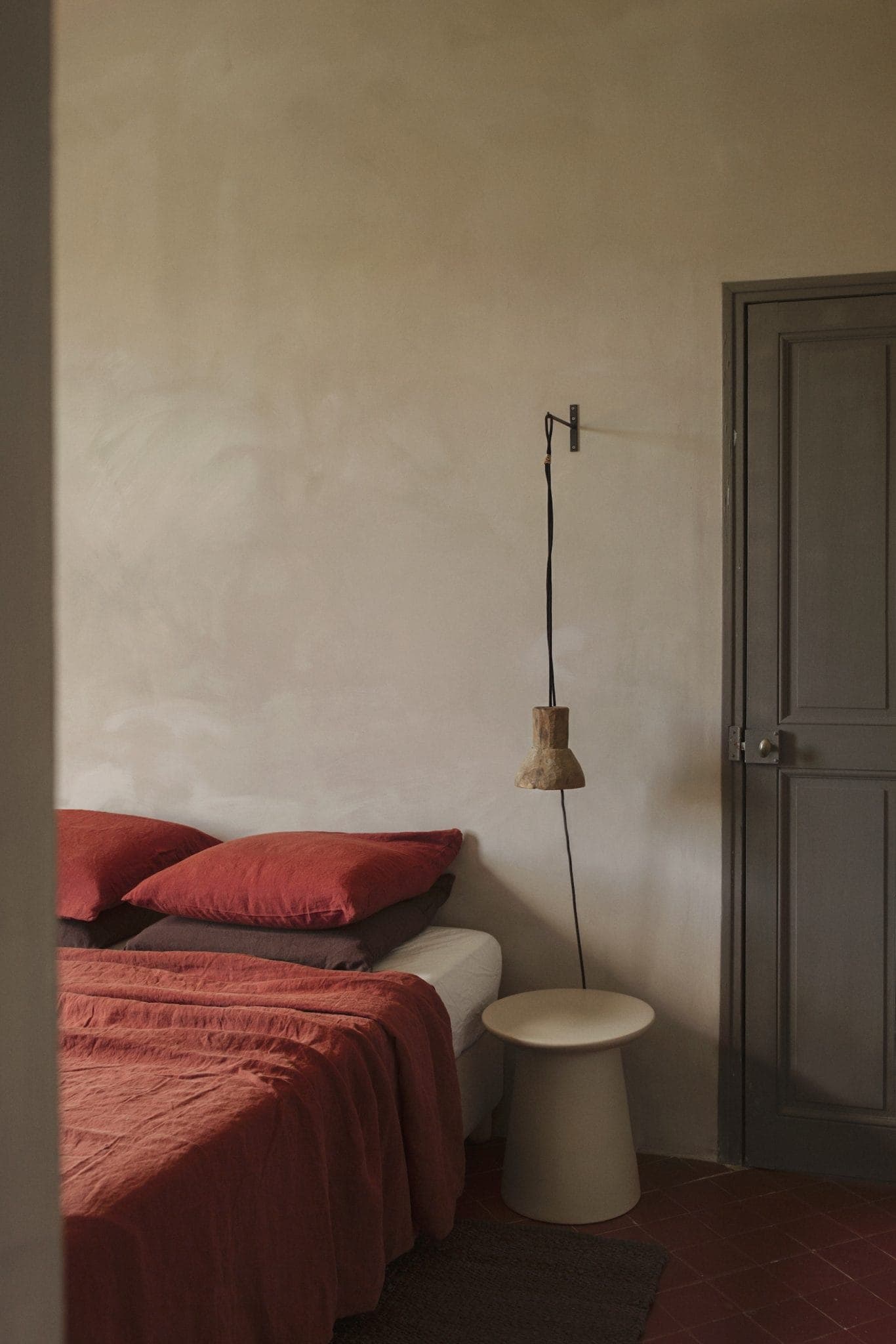 A minimalist bedroom with a bed covered in rust-colored sheets and pillows. A small round side table with a hanging lamp above is next to the bed. The walls are neutral, and there is a closed gray door to the right.