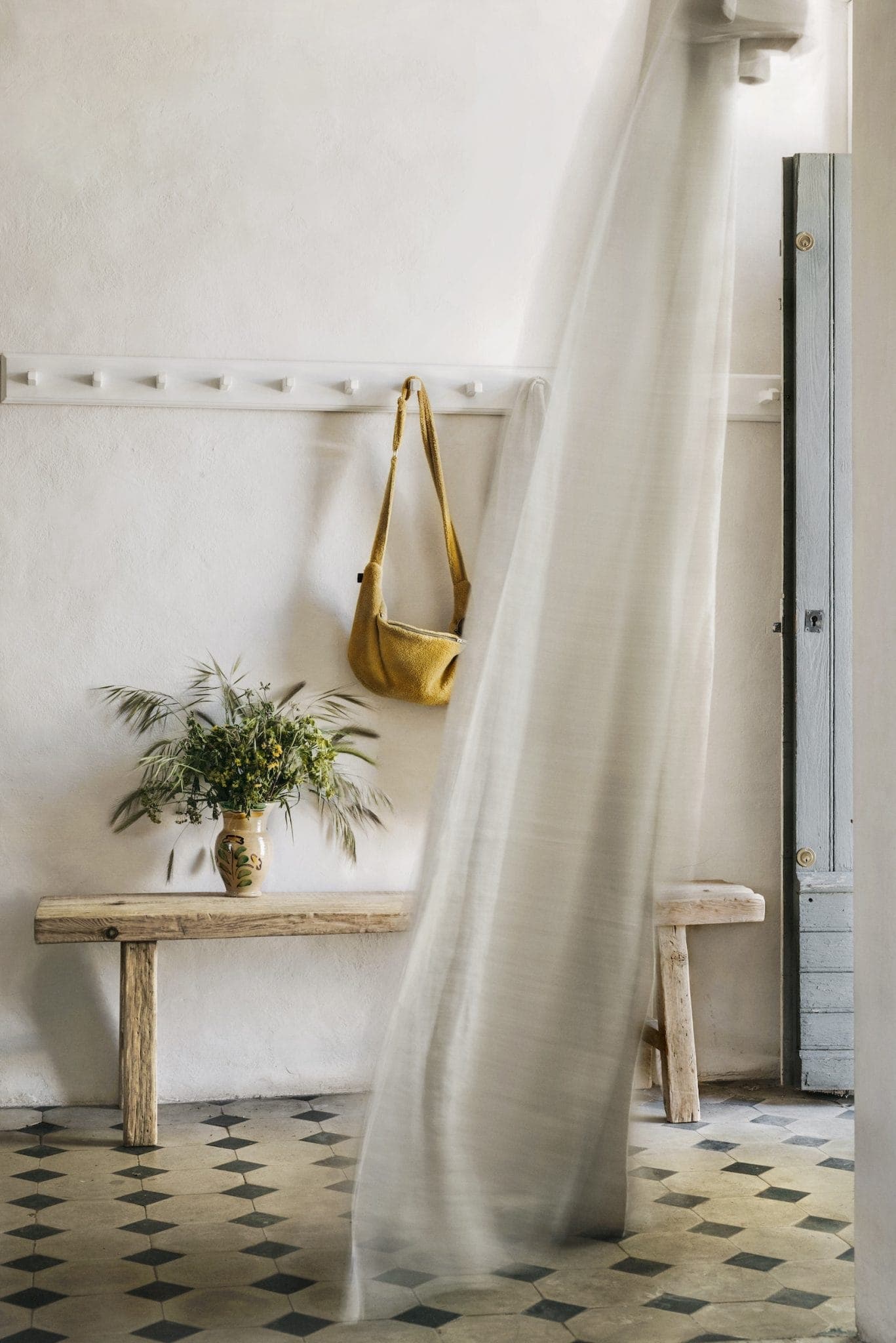 Bright, airy room with a light curtain billowing. A rustic wooden bench holds a vase of flowers. A mustard-colored bag hangs on a wall hook. The floor has geometric tiles, and a partially open door allows sunlight to stream in.