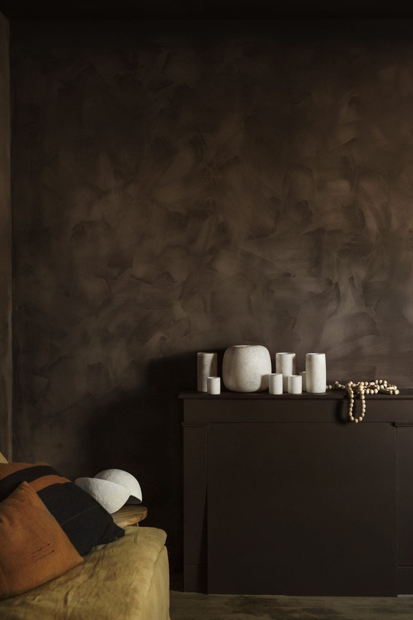 Dimly lit room with textured dark brown walls. A dark console holds several white ceramic vases of various shapes and sizes, alongside a string of beads. A beige couch features brown and white pillows. Minimalist and earthy decor.