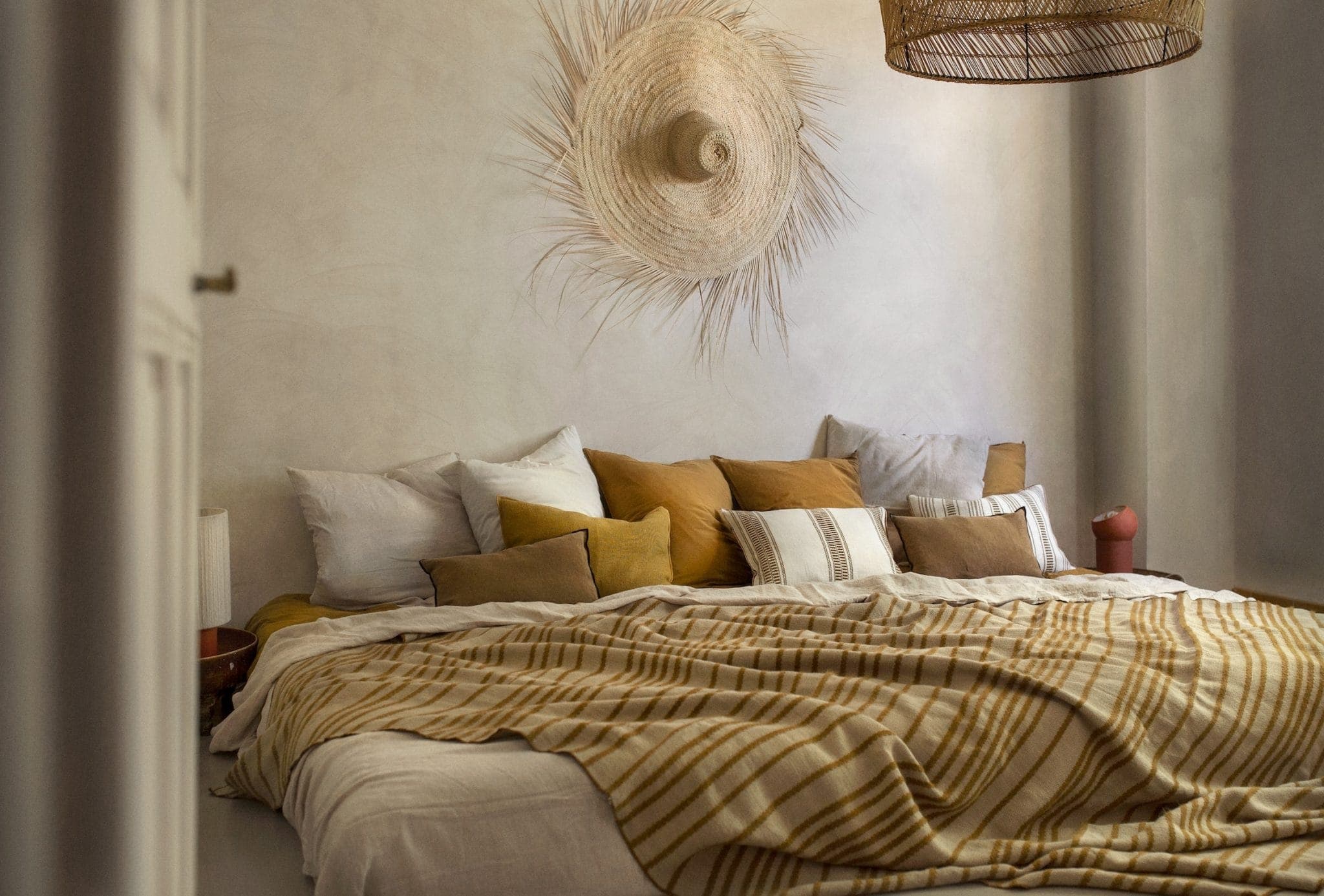A cozy bedroom features a made bed with white and amber pillows, covered with a checkered blanket. A straw hat decorates the wall, and a wicker lampshade hangs from the ceiling, creating a warm and inviting atmosphere.