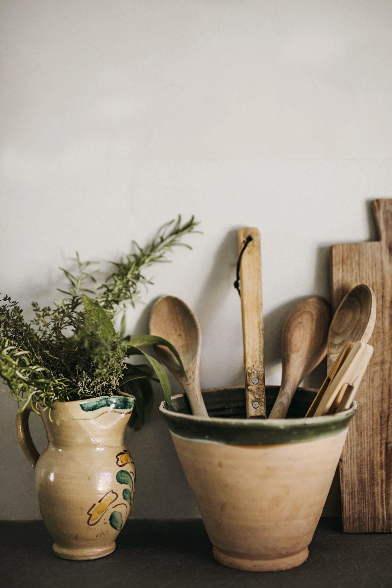 Ceramic pots on a shelf; one holds fresh herbs like rosemary and thyme, and the other contains wooden kitchen utensils. A few cutting boards are leaned vertically against the wall behind them.