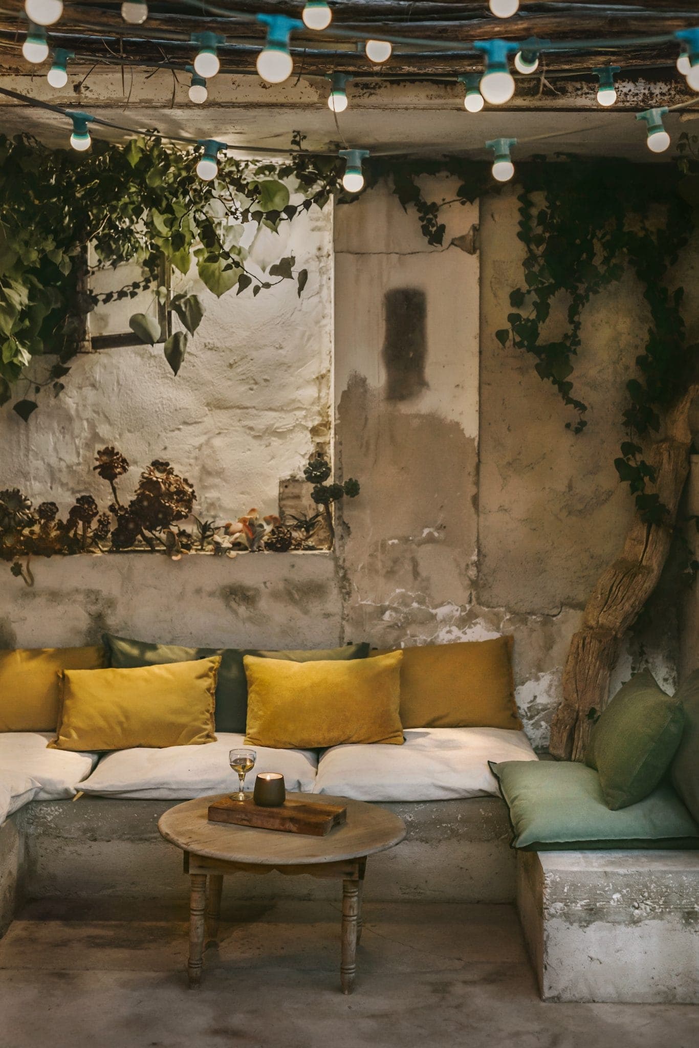 Cozy seating area with a rustic vibe, featuring yellow and green cushions on a concrete bench. A small wooden table holds a drink and a candle. Overhead, string lights and greenery create a warm, inviting atmosphere.