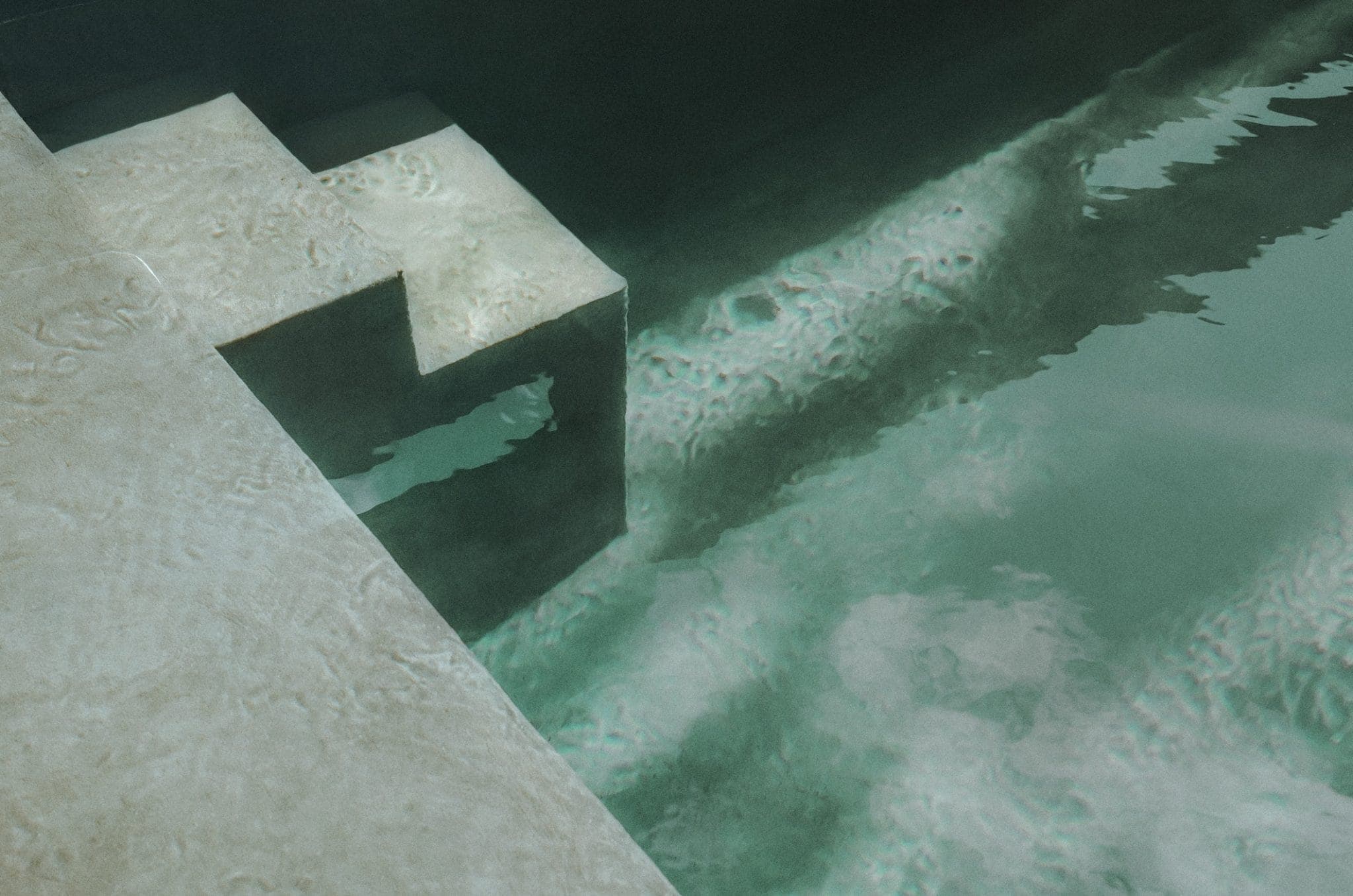 A serene swimming pool with rippling water reflecting light. White stone steps lead into the turquoise water, surrounded by dark walls, creating a tranquil and inviting atmosphere.