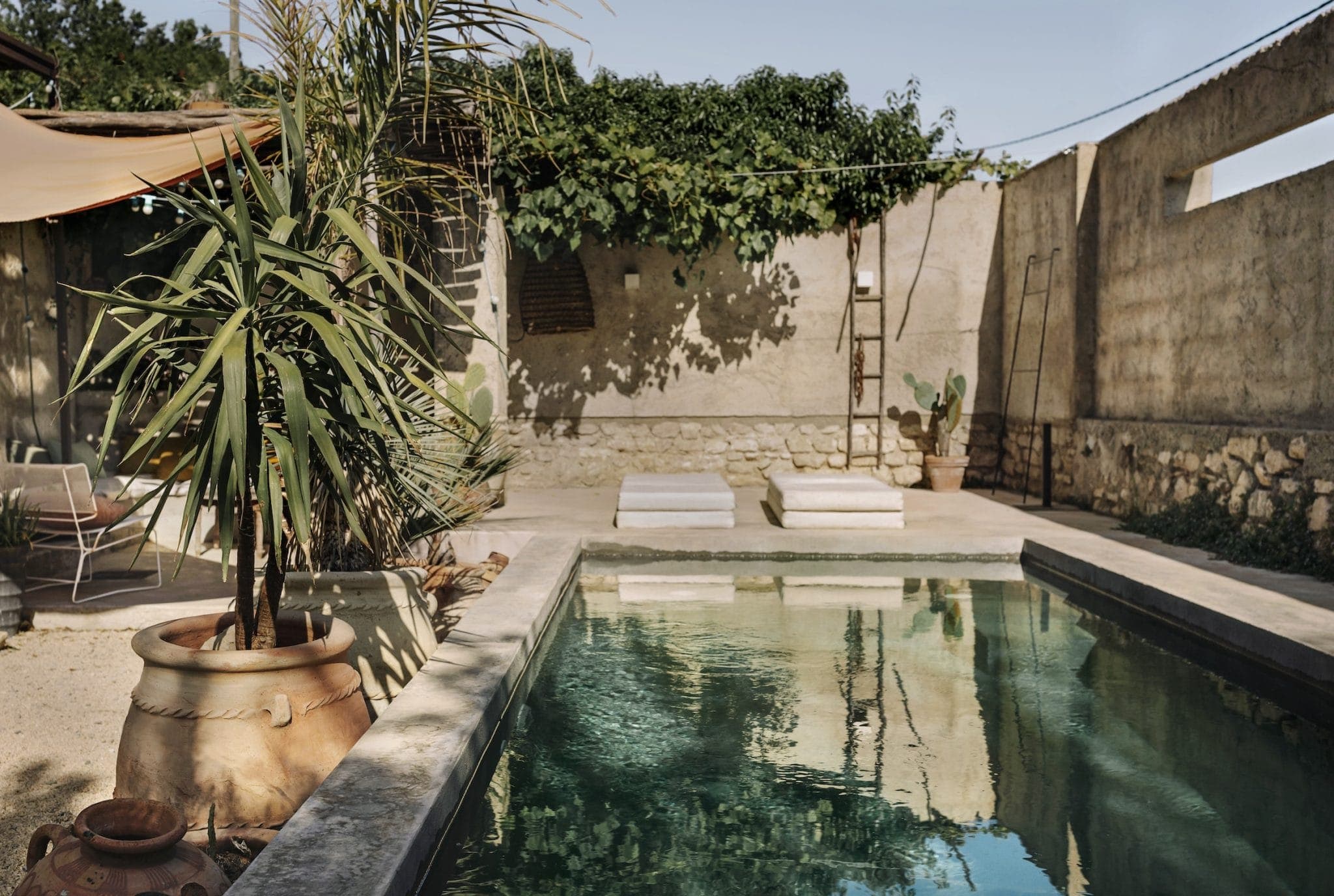 A serene outdoor setting features a rectangular pool with clear water reflecting surrounding walls and greenery. There are potted plants, cushioned seating, and a rustic stone and stucco wall, creating a tranquil, Mediterranean ambiance.