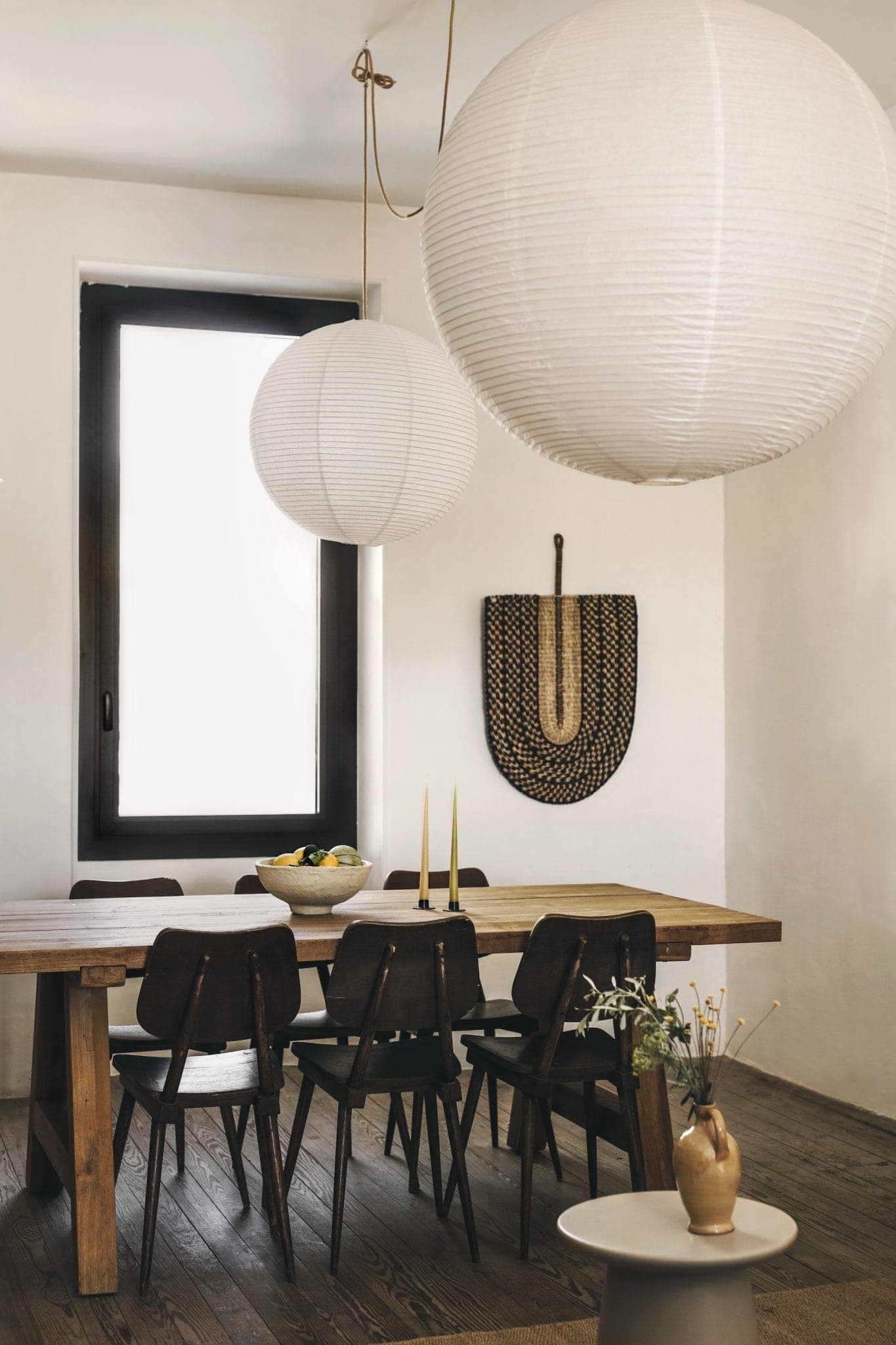 A dining room with a wooden table and six dark chairs. Two large white paper lanterns hang from the ceiling. A decorative woven piece is on the wall. A bowl of fruit and tall candles are on the table. A small round side table holds a vase.