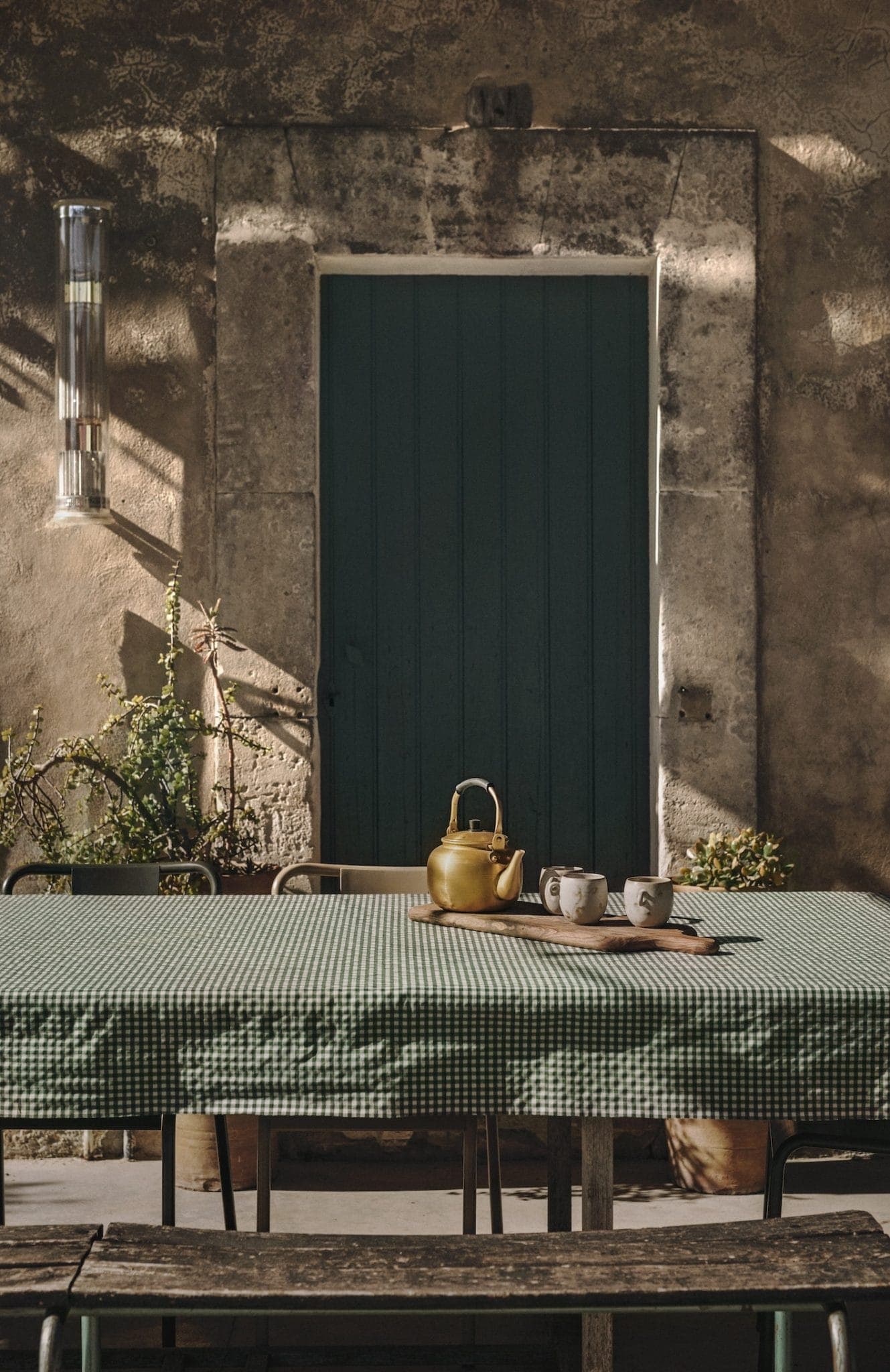 A sunlit outdoor table with a green checkered cloth holds a brass teapot and two mugs on a tray. Behind it, an old stone wall features a closed teal door. Sunlight casts shadows on the wall.