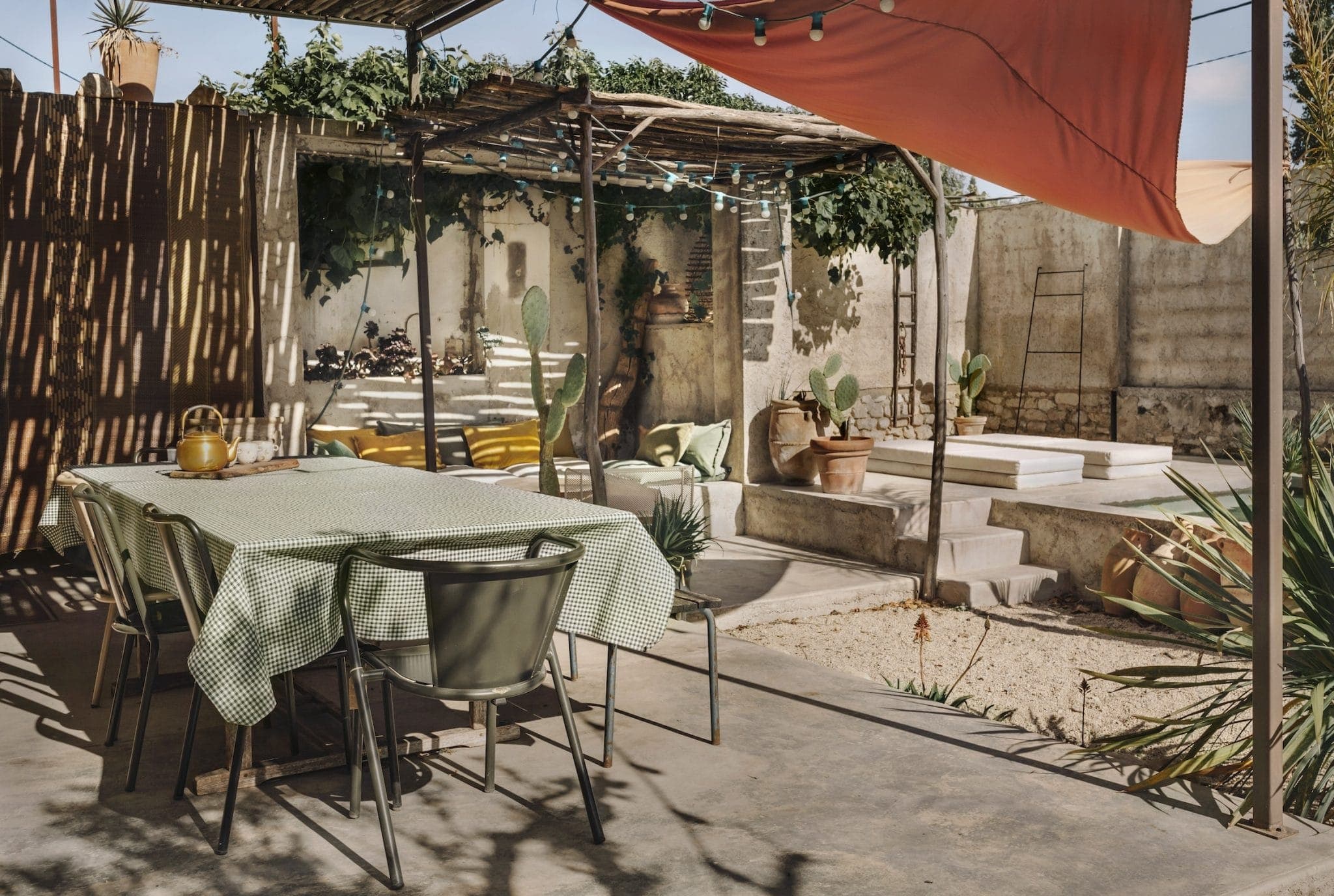 Outdoor patio dining area with a table covered by a green checkered cloth, surrounded by chairs. Overhead, a pergola with hanging lights offers shade. Steps lead to a raised section with cacti and potted plants. A red canopy adds color on the right.