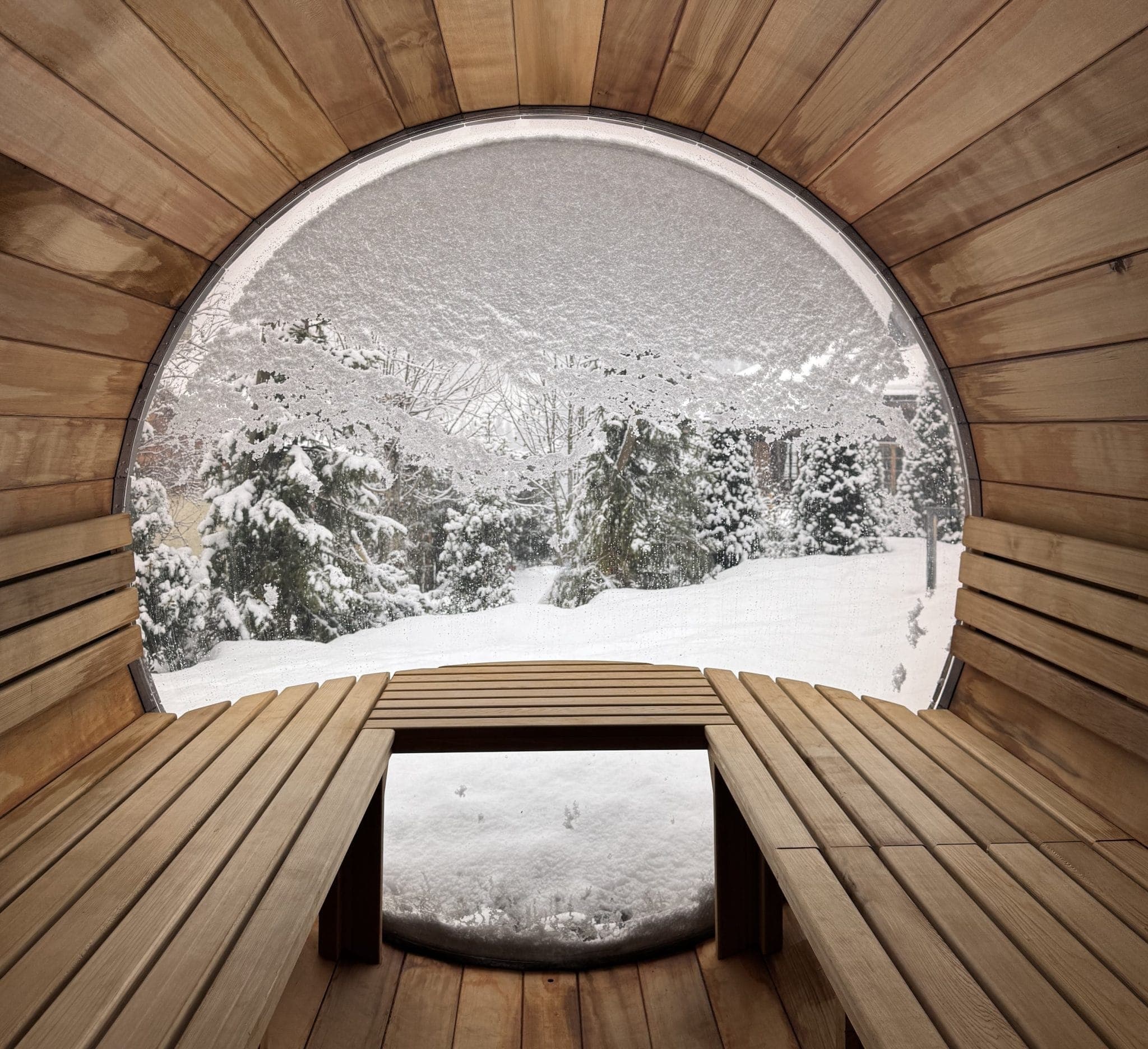 Interior of a circular wooden sauna with benches on either side. The large window shows a snowy landscape with trees and a thick layer of snow outside, creating a cozy and serene winter scene.