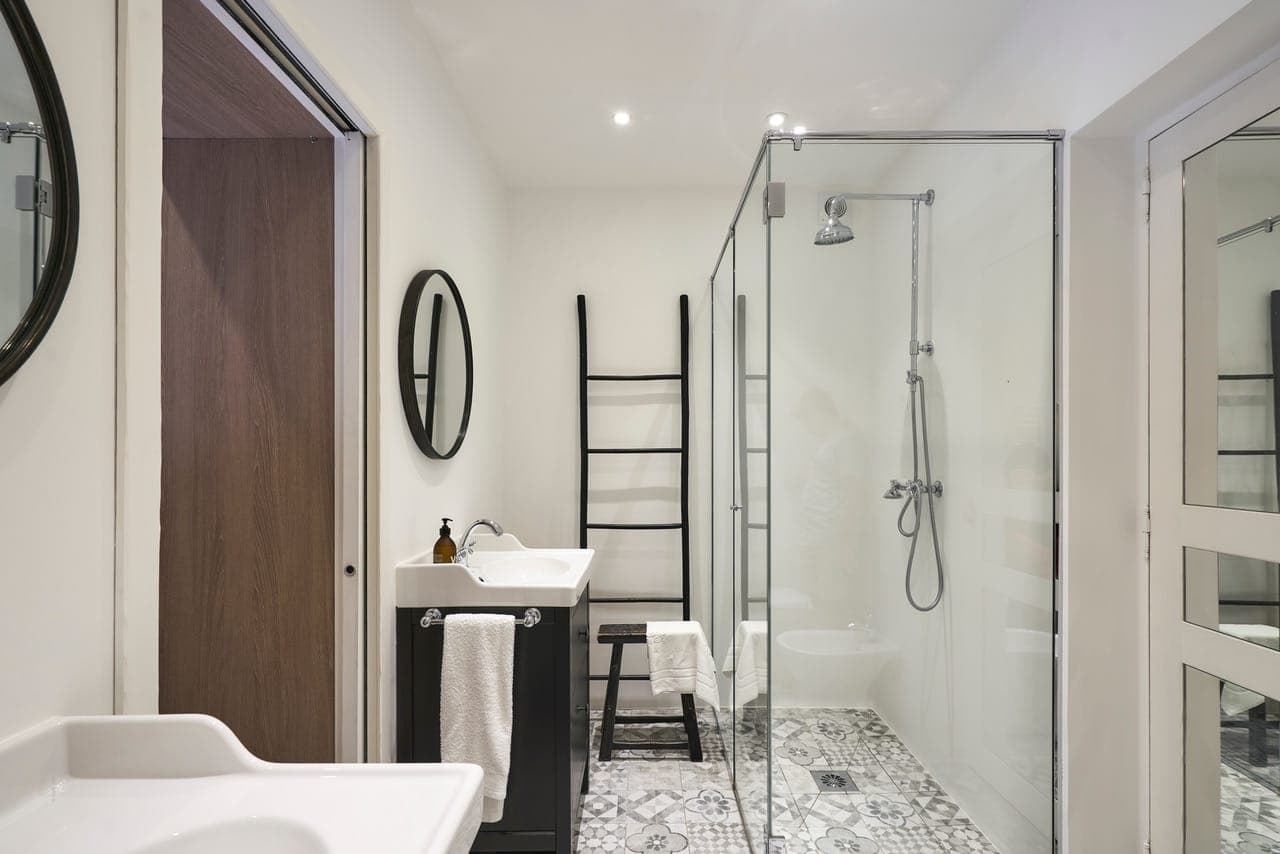 Une salle de bain moderne avec une douche vitrée, un carrelage à motifs, un lavabo mural avec un meuble noir et un miroir rond. Il y a une porte en bois à gauche et un porte-serviettes noir de style échelle. Les murs blancs et les plafonniers créent une atmosphère lumineuse.
