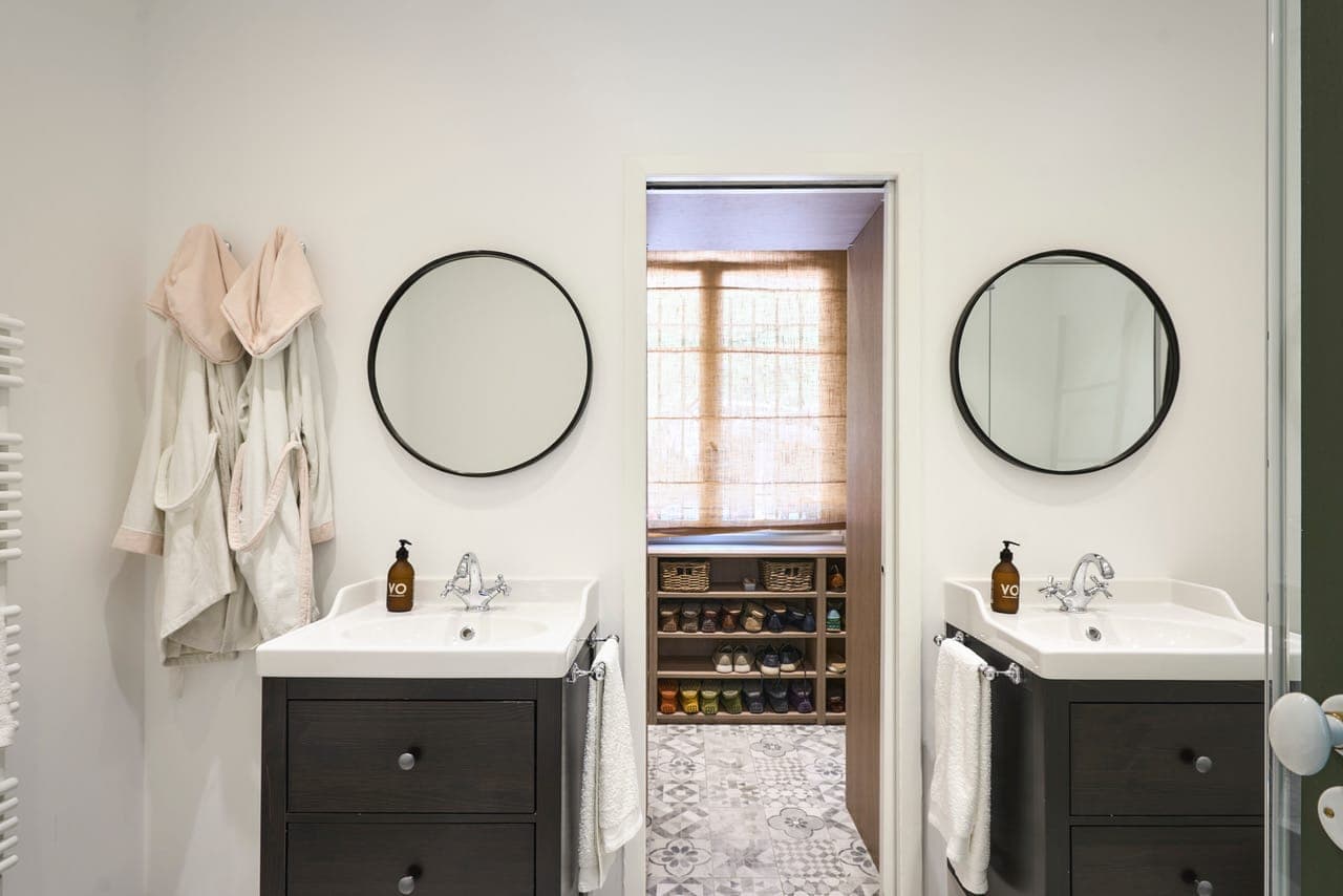 Une salle de bain lumineuse avec deux miroirs ronds au-dessus de vanités noires, chacune avec des lavabos blancs et des distributeurs de savon marron. Des serviettes blanches sont suspendues à côté d'eux. Les peignoirs sont sur la gauche. Une porte s'ouvre sur une pièce ensoleillée avec des étagères à chaussures et un sol en carrelage complexe.