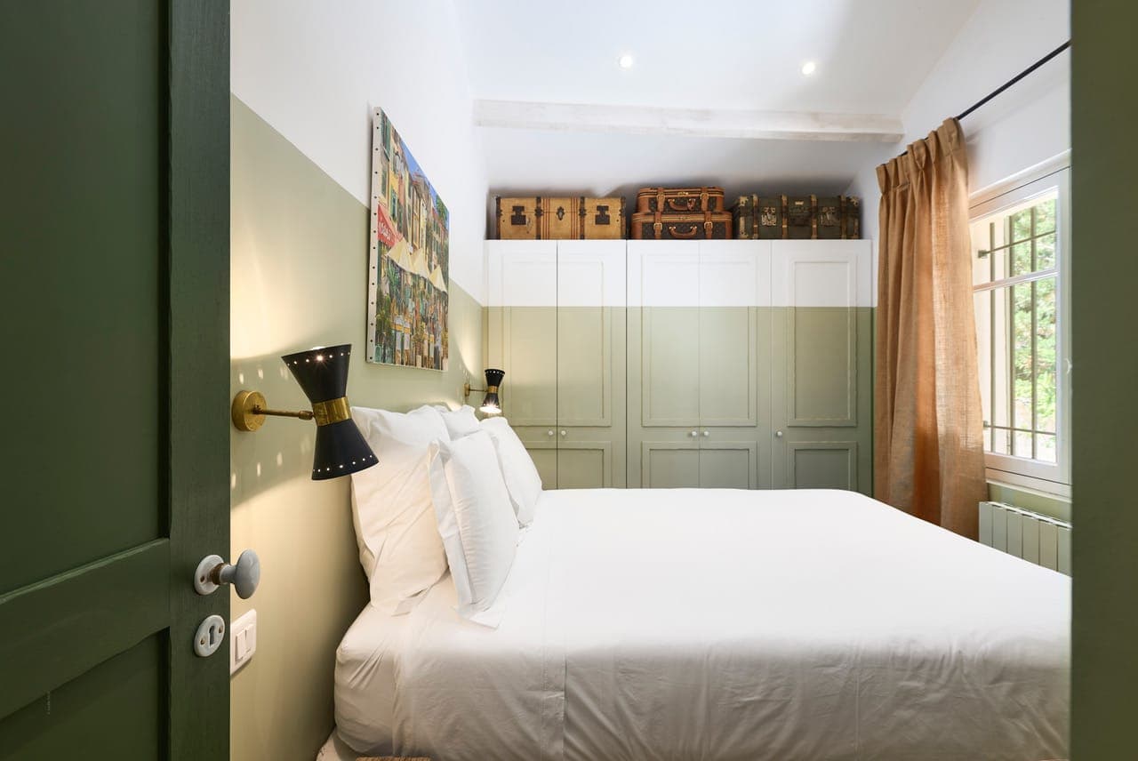 Une chambre confortable avec un grand lit blanc, des murs verts à moitié peints et des armoires vertes encastrées. Des valises vintage sont placées au-dessus des armoires. Une grande fenêtre avec des rideaux beiges laisse entrer la lumière naturelle. Des appliques murales rétro sont placées près du lit.