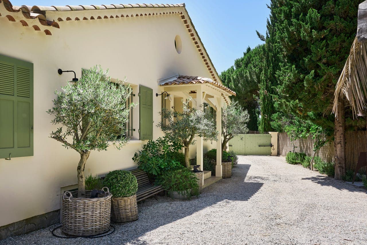 Une charmante maison aux murs beige clair et aux volets verts, entourée de plantes en pot et de petits arbres. Un chemin de gravier mène à une entrée fermée. Un ciel bleu clair et de grands arbres encadrent la scène, créant une atmosphère paisible.
