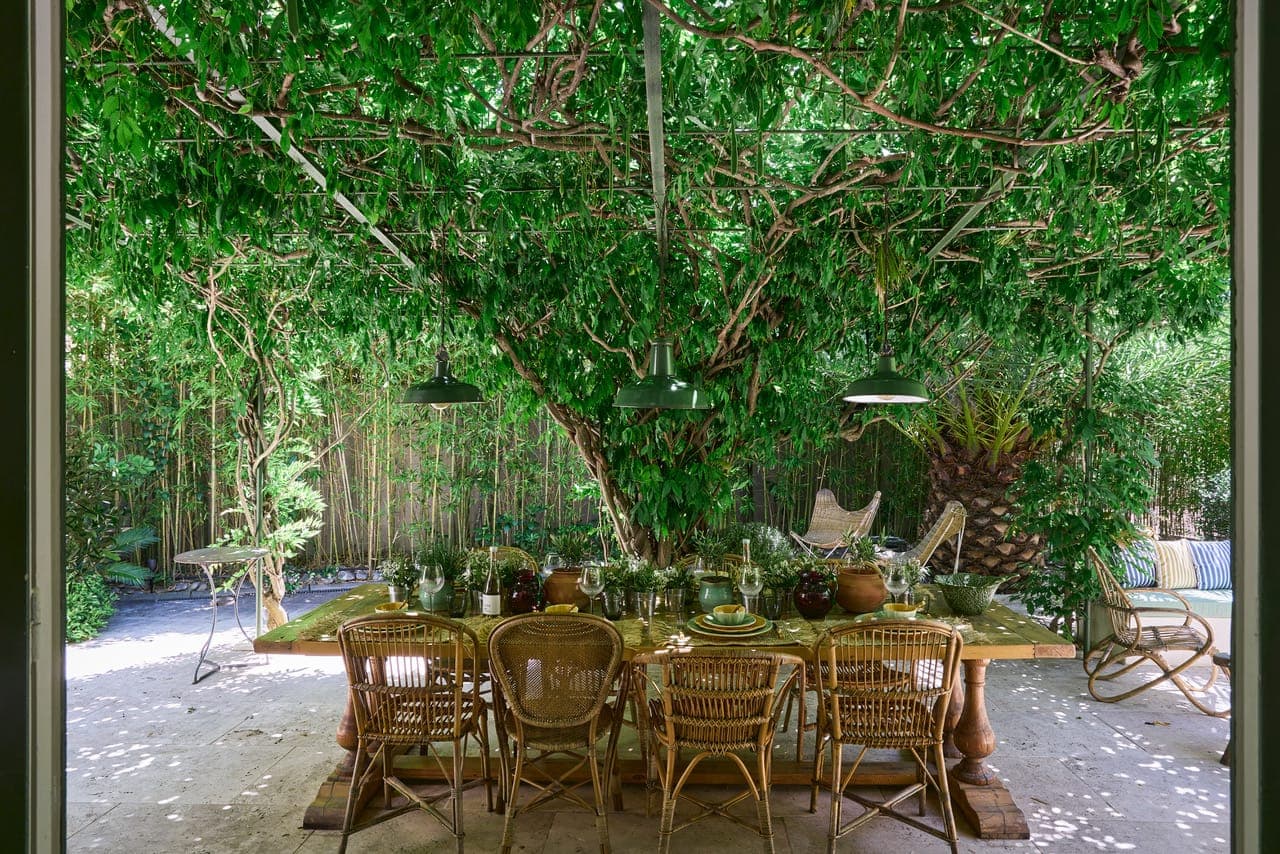 A lush garden setting with a long wooden dining table surrounded by wicker chairs. The table is set with plates and glasses under a canopy of dense green foliage. Hanging overhead are three green lamps. The background features bamboo and outdoor furniture.