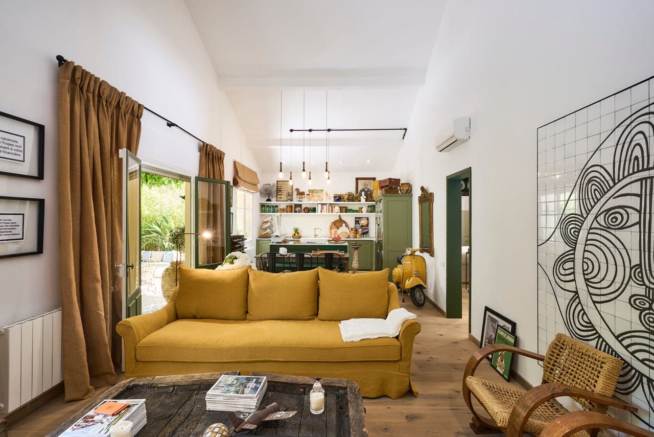 Un salon confortable avec un canapé jaune moutarde face à une table basse en bois rustique. La pièce présente une œuvre d'art à motif quadrillé, de grandes fenêtres et un coin repas visible avec un scooter garé à l'intérieur. La décoration comprend des chaises en osier et des œuvres d'art encadrées.