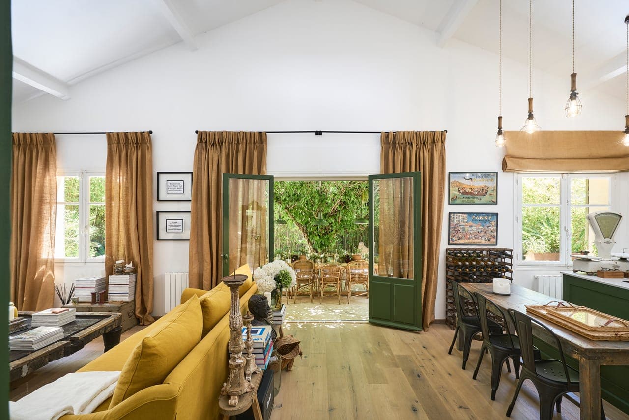 Un salon lumineux avec un canapé jaune, des tables en bois et une porte ouverte menant à un patio extérieur. La pièce présente une décoration rustique, de grandes fenêtres avec des rideaux marron, du parquet et des suspensions.