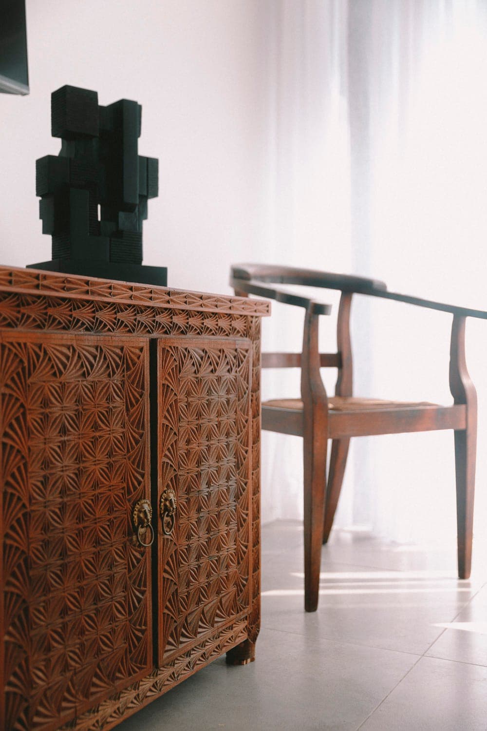 A wooden cabinet with intricate carvings and elaborate handles stands next to a modern, curved wooden chair. A black geometric sculpture rests on top. Soft light filters through transparent curtains, creating a serene atmosphere.