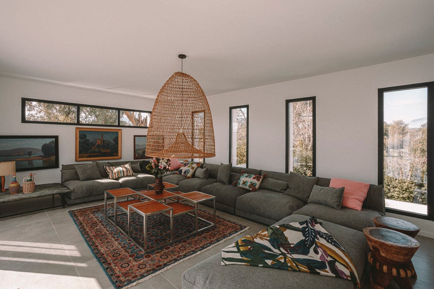 A spacious living room with large windows features a wide gray corner sofa adorned with colorful cushions. A woven pendant hangs above a coffee table set on a patterned rug. Wall art and natural light add to the warm atmosphere.