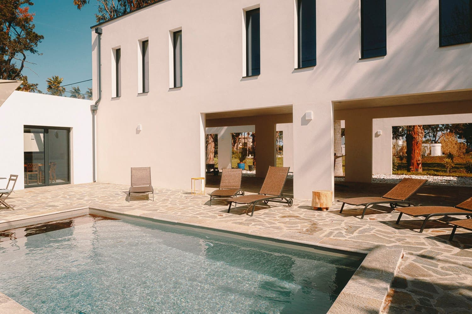 Modern villa exterior with a rectangular swimming pool surrounded by stone tiles. Several lounge chairs are placed around the pool. The building features large windows and a covered patio area, set against a clear blue sky.