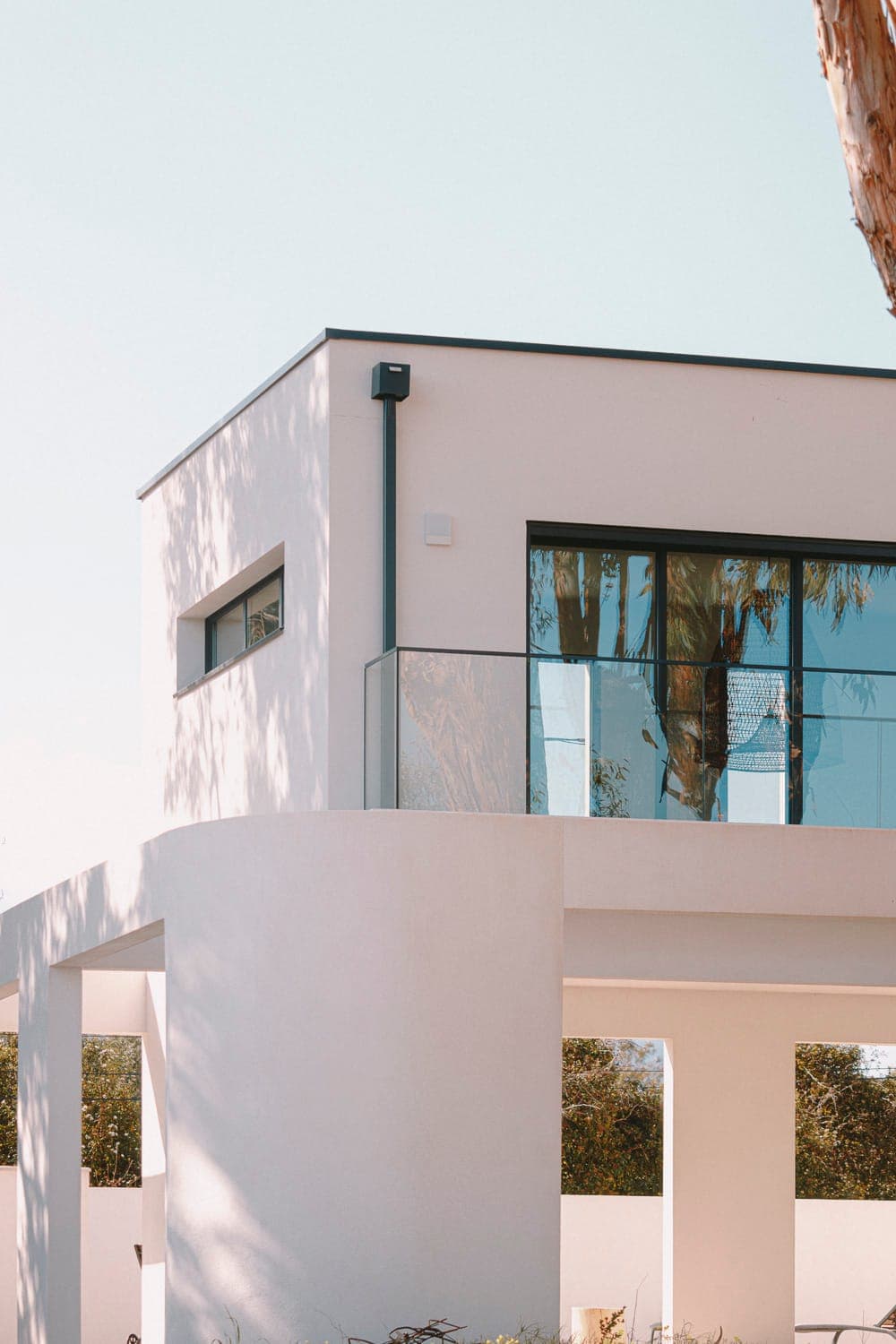 A modern, minimalist house with white walls and large glass windows. The building has a flat roof and a spacious balcony with a glass balustrade. The sky is clear and the trees cast soft shadows on the building.