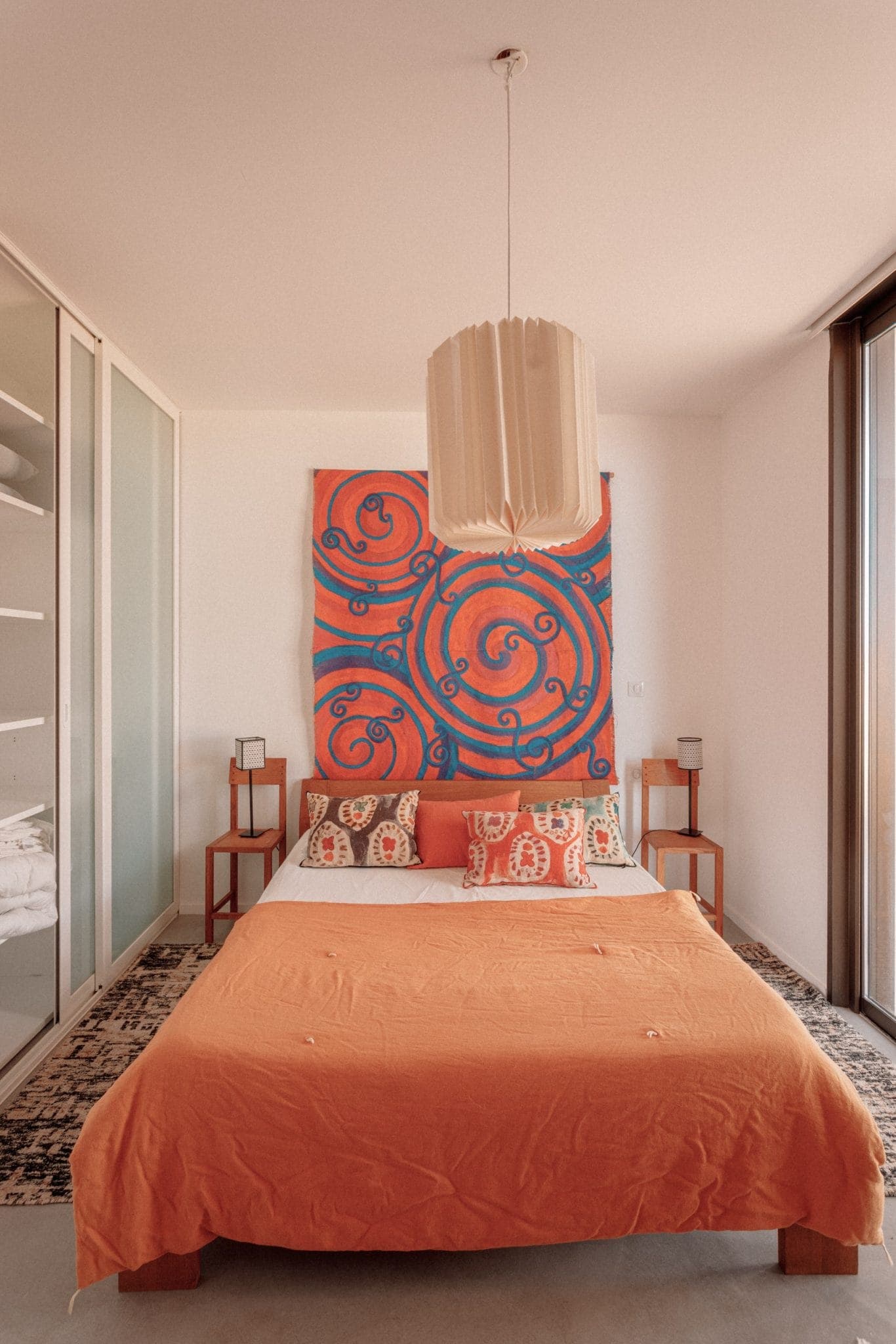 A comfortable room with a bed with an orange comforter and patterned pillows. A large red and blue abstract painting hangs above the bed. Wooden bedside tables with small lamps are arranged on either side, and a modern hanging lamp hangs from the ceiling.