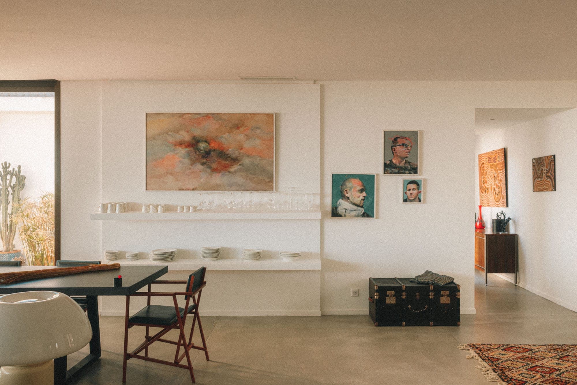 A modern living room with a white wall adorned with abstract paintings and portraits. A black dining table with a red chair and white bowls is in the foreground. A large window brings natural light into this minimalist space.