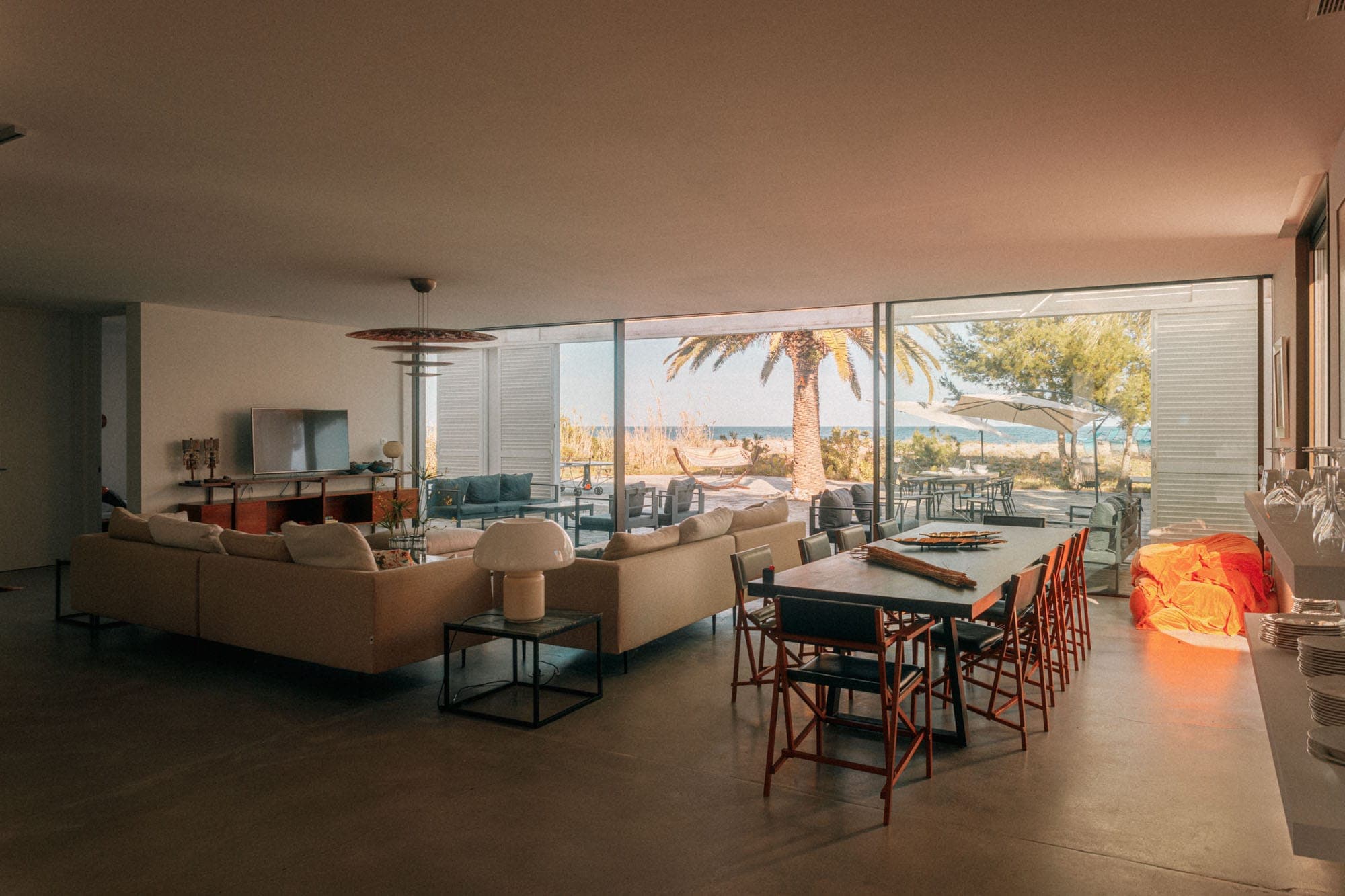 A spacious, modern living and dining room with large windows offering views of the beach. The room features a beige corner sofa, a dining table with wooden chairs and an outdoor patio with table and parasol, surrounded by palm trees.