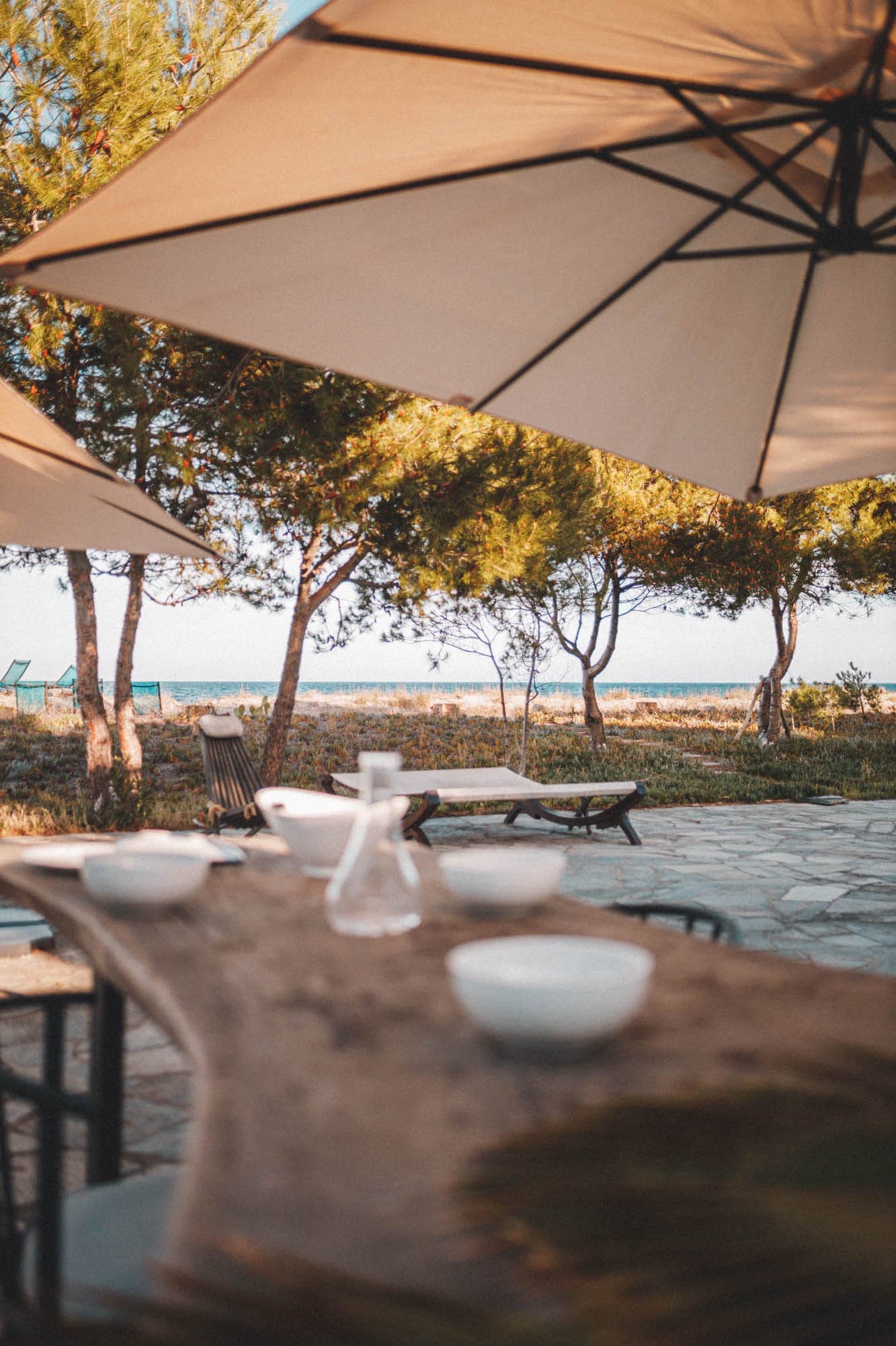 A wooden table outside with white bowls and a glass pitcher under large beige parasols. In the background, trees and a glimpse of the sea suggest a seaside setting. Sunlight filters through, creating a warm atmosphere.